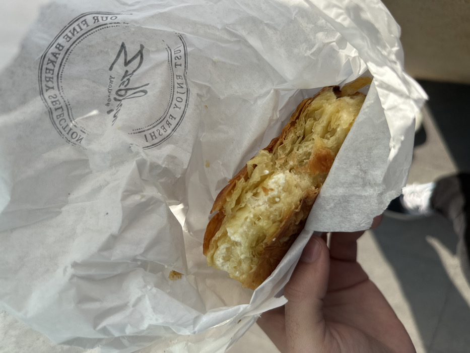 The image shows a person's hand holding a partially eaten pastry inside a white paper bag under bright sunlight. The pastry features golden-brown, flaky layers of phyllo dough with a visible white, creamy cheese filling, resembling a spanakopita or tiropita.
The white paper bag has a circular logo printed on it, with text that appears mirrored or reversed. Along the left arc, the text "OUR FINE BAKERY SELECTION" is visible, and along the right arc, "JUST ENJOY FRESH!" can be read. A stylized central letter, possibly an "M" for "Mela," with "Bakery" below it, is also discernible.
The scene is outdoors, likely on a sunny day given the strong shadows cast on the ground. The shadow of a person's legs and shoes is visible on a light-colored paved surface in the background, suggesting an outdoor location in Metamorfosi, Greece. The focus is on the food item and the paper bag, capturing the moment of consuming the pastry.
