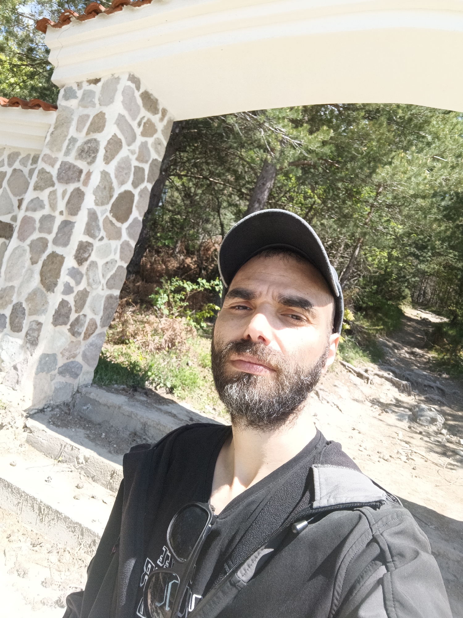 A selfie taken by a man with a beard wearing a gray baseball cap and a black jacket with sunglasses hanging from the neck area. He is standing under a stone archway with a white top and red tiles on top of that. The archway is situated at the entrance to a dirt path leading into a wooded area. The location is Sini Vrah, Bulgaria.