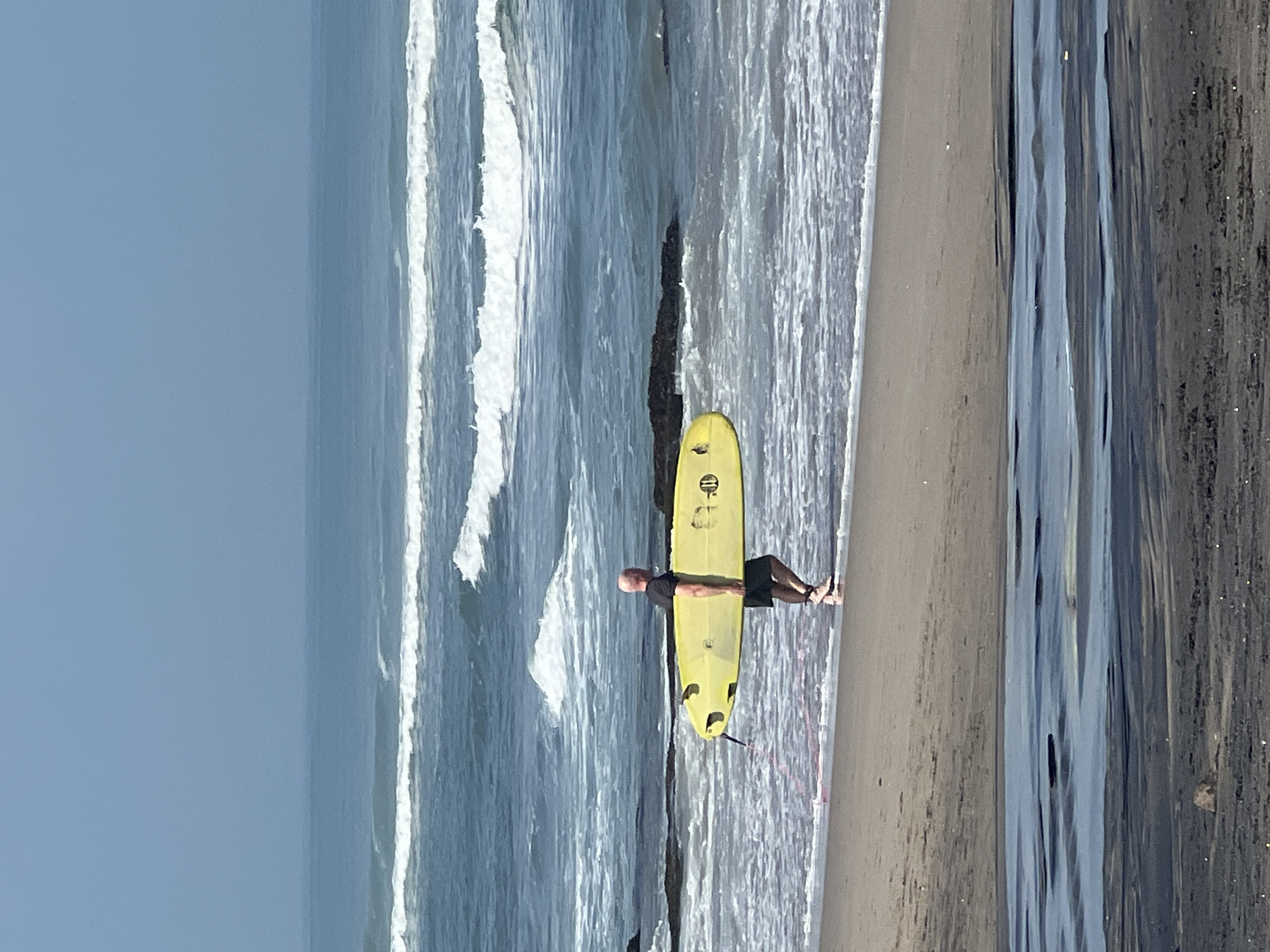 A bald man, wearing a dark t-shirt and black shorts, carries a bright yellow surfboard with a pink leash under his right arm. He is walking along the wet sand near the water's edge on a beach. The ocean has moderate waves breaking, with white foam visible. In the background, the sea extends to the horizon under a hazy blue sky. The beach appears to be composed of sand and dark wet areas closer to the foreground, suggesting a tidal zone or shallow water. The scene is likely located at a beach in North Kuta, Indonesia, given the prevalence of surfing and similar coastal landscapes in the region.
