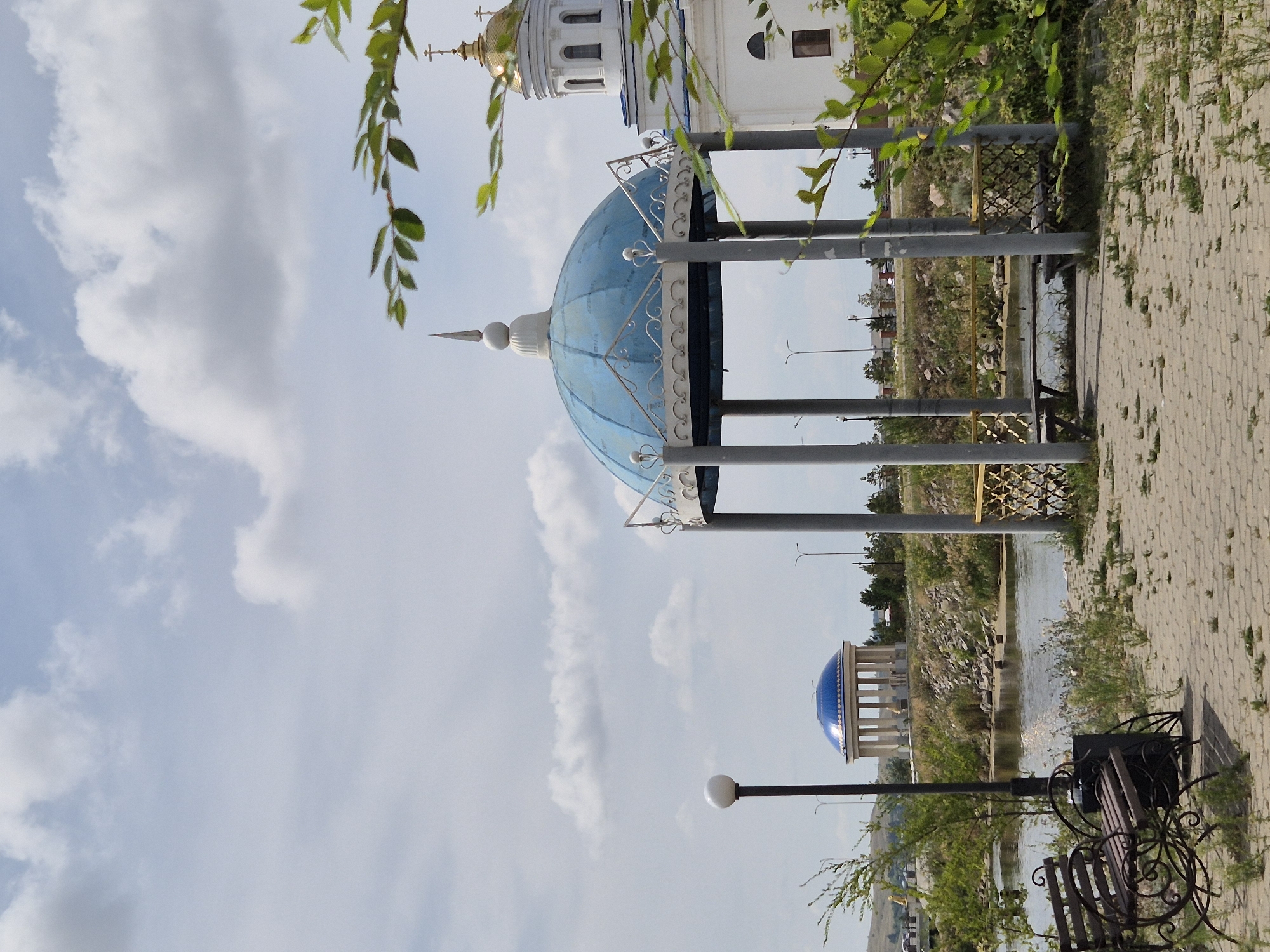 The image depicts a park-like setting in Kokshetau, Kazakhstan, with a prominent blue-domed gazebo in the foreground. The gazebo is supported by grey pillars and features ornate white detailing. Behind it, to the right, a white building with a golden dome, likely a church, is partially visible. Across a body of water, a rotunda with a blue dome and classical columns stands on a hill. In the foreground, a bench made of dark metal and wood sits on a paved walkway. The environment is characterized by a cloudy sky, lush green vegetation, and a calm body of water. No people are visible in the scene.