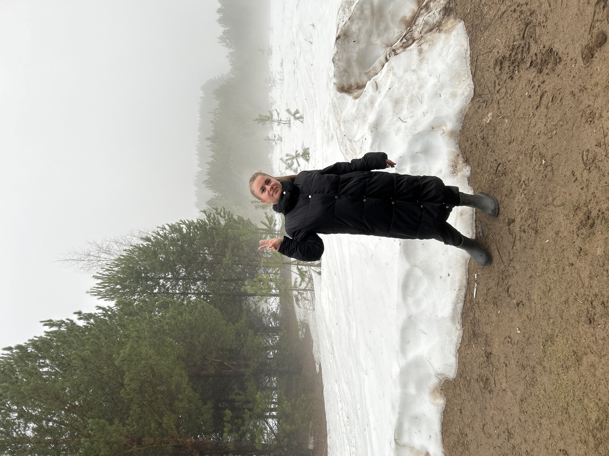 The image shows a person standing on a snowy and muddy terrain, likely near a forest. The person is wearing a long black puffer coat and boots, and is making a peace sign with their hand. The background features a misty forest and patches of snow. Given the information provided and the appearance of the environment, this image was likely taken in the outskirts of Chaikovsky, Russia where forests, snow, and mud is common.