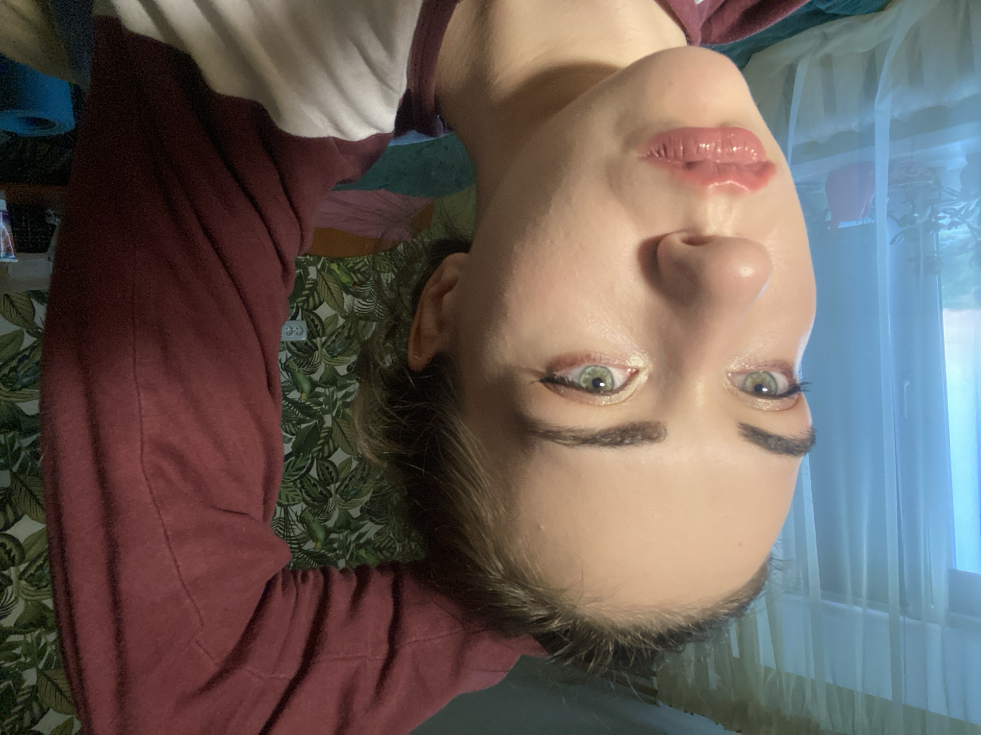 The image shows a close-up, profile view of a young woman's face, her right arm raised and resting behind her head. She is wearing a maroon sweatshirt. Her makeup includes eye shadow and lip gloss. She appears to be indoors, with sheer curtains and a window visible in the background to her left, and a patterned wallpaper behind her. A bed or sofa is visible below her right arm. There is no indication of the city of Zavodoukovsk, Russia, in the image.