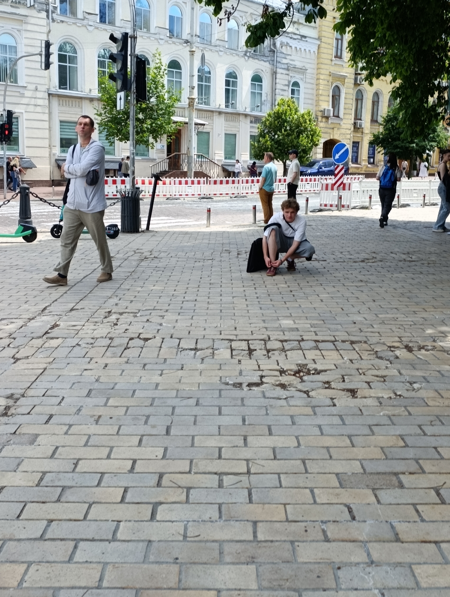 This media file captures an outdoor scene in Kyiv, Ukraine. The foreground is dominated by a cobblestone walkway, showing signs of wear and tear with some stones missing or uneven. In the middle ground, several people are present. To the left, a man stands with his arms crossed, wearing a light-colored hooded jacket and beige pants. Next to him, a green electric scooter is parked. Further into the middle ground, a young man is crouched down, tying his shoelaces, with a black bag beside him. Other individuals can be seen walking in the background.
Behind the people, a street scene unfolds. There are buildings with architectural details, including arched windows and ornate facades. A traffic light stands prominently near the man in the hooded jacket. Construction or renovation barriers, marked with red and white, are visible in front of some buildings. A blue directional sign with a white arrow is also present. The scene appears to be set during the daytime, with bright lighting suggesting fair weather. The overall atmosphere suggests a normal day in a city environment, with people going about their activities on a public street.
Visible text includes "CREDIT AGRICOLE" on the building to the left, and a license plate number "T-369" on the electric scooter.