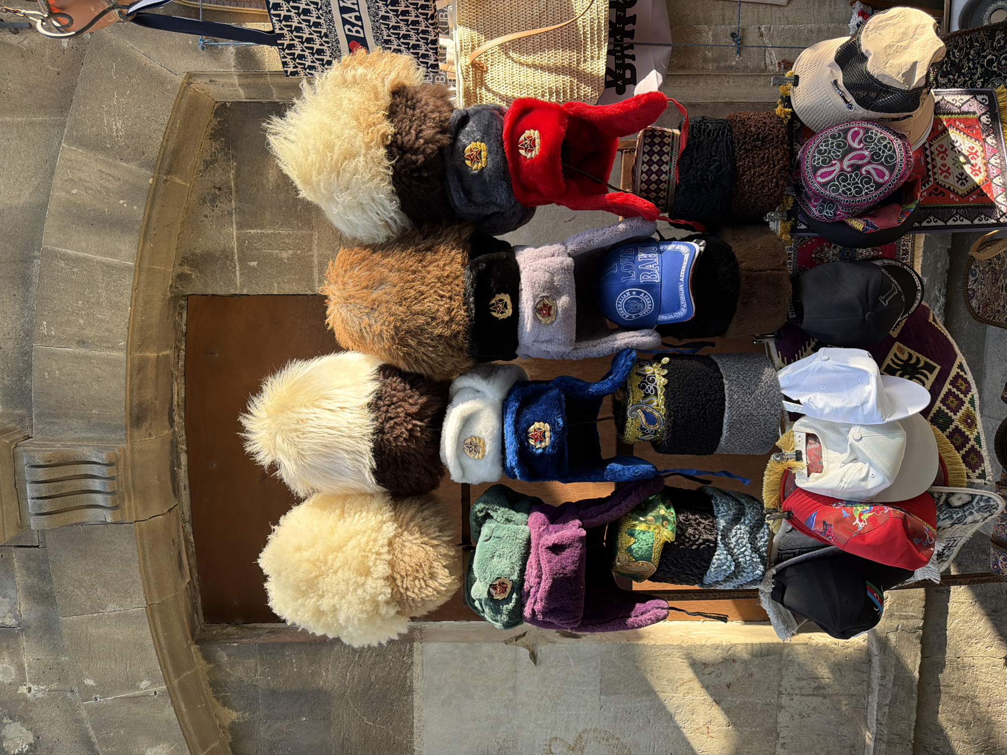 This is a medium shot, eye-level, outdoor photograph of a hat stall located in Sabail Raion, Azerbaijan, likely near the Old City or a tourist area. The stall displays a variety of hats, including traditional fur hats with ear flaps, often referred to as ushanka hats, and baseball caps. The hats are arranged in stacks and hung from metal hooks. The background consists of an old stone building with an arched doorway. Sunlight casts shadows across the scene, indicating it is daytime. There are no people visible in the image. The hats themselves are diverse in color and texture, with some featuring embroidery and badges. Some of the fur hats appear to be made of sheepskin or similar material. The overall impression is one of a bustling marketplace or souvenir vendor.