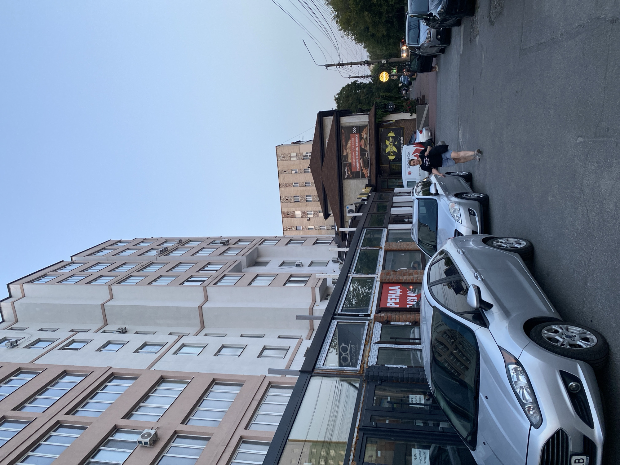 This is a photograph taken at dusk on an urban street in Vinnytsia, Ukraine. The scene features a multi-story building with a light pink facade on the left and a white and beige facade on the right, featuring numerous windows. In front of these buildings are commercial establishments with large glass windows and black framing. A red sign with Cyrillic text, "РЕHДА", is visible on one of the storefronts. Several silver cars are parked along the side of the street, including a Ford Fiesta in the foreground. A woman in a black t-shirt and denim shorts is walking away from the viewer on the right side of the street, carrying a black bag. Further down the street, more cars are parked, and a street lamp with a circular, illuminated fixture is visible. The sky is a clear, pale blue, indicating the end of daylight. The overall impression is of a quiet evening on a commercial street in a Ukrainian city.