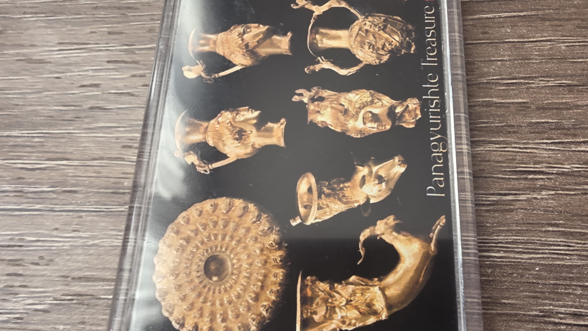 The image shows a photograph of a display featuring artifacts from the Panagyurishte Treasure. The treasure consists of golden Thracian rhyta (drinking vessels), a phiale (bowl), and an amphora. The artifacts are prominently displayed against a black background. The text "Panagyurishte Treasure" is partially visible on the right. The setting appears to be indoors. The Panagyurishte Treasure is associated with the city of Panagyurishte, Bulgaria, not Blagoevgrad.