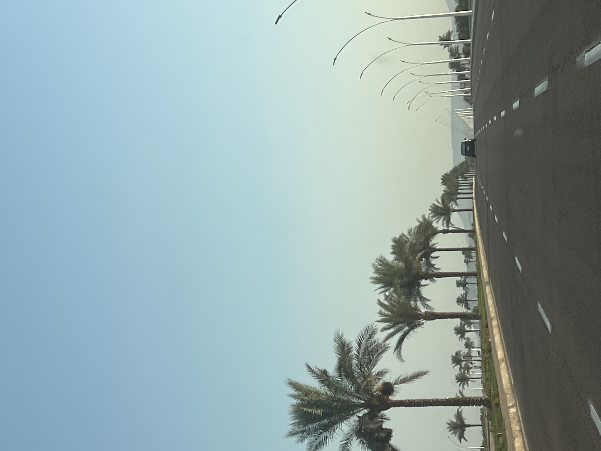 The image depicts a road in Sharm El Sheikh, Egypt, lined with palm trees and modern streetlights. The road is paved and has white dashed lines indicating lanes. A dark-colored car is visible driving away from the camera down the road. The sky is a clear, pale blue, transitioning to a lighter hue near the horizon. In the distance, faint outlines of hills or mountains can be seen. The overall scene suggests a well-maintained urban or coastal area.