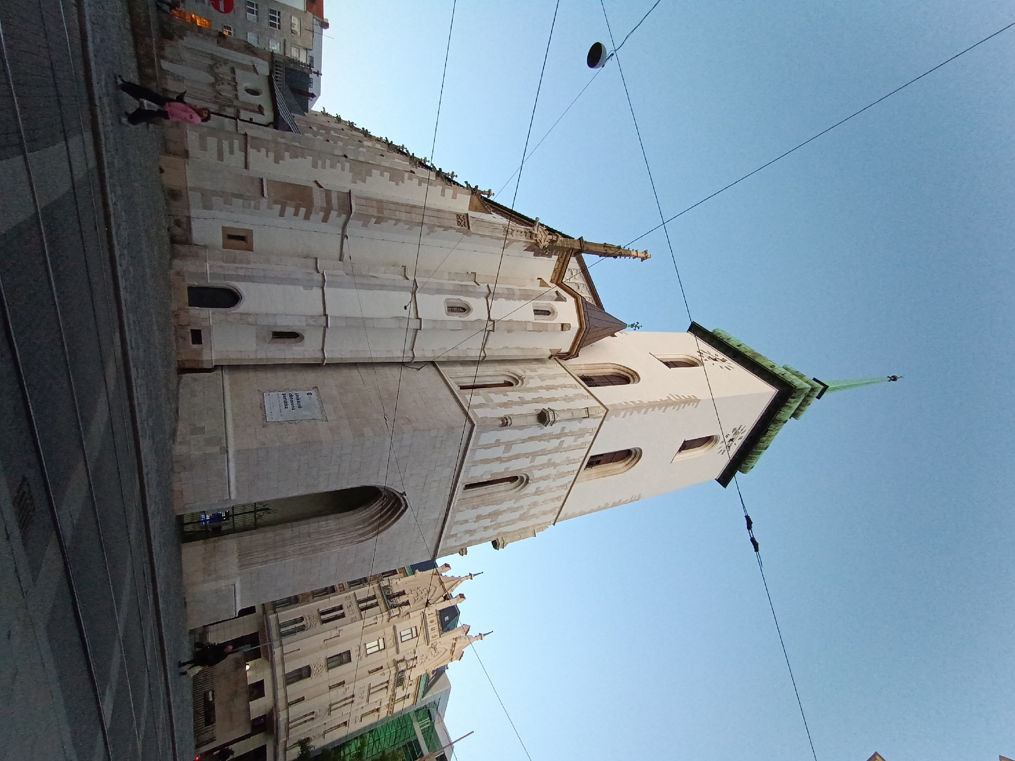 The image shows the St. Peter and Paul Cathedral, a prominent landmark in Brno, Czechia, viewed from a low angle. The cathedral's Gothic architecture is evident, with its tall, white facade, arched windows, and a green spire. A clock face is visible on the tower. The sky is clear and blue. In the foreground, tram tracks run across a cobblestone street. A few individuals are walking on the sidewalk. To the right of the cathedral, another building with ornate architecture and a prominent date "1905" is visible. Overhead, utility wires and a street lamp are present, suggesting an urban setting.