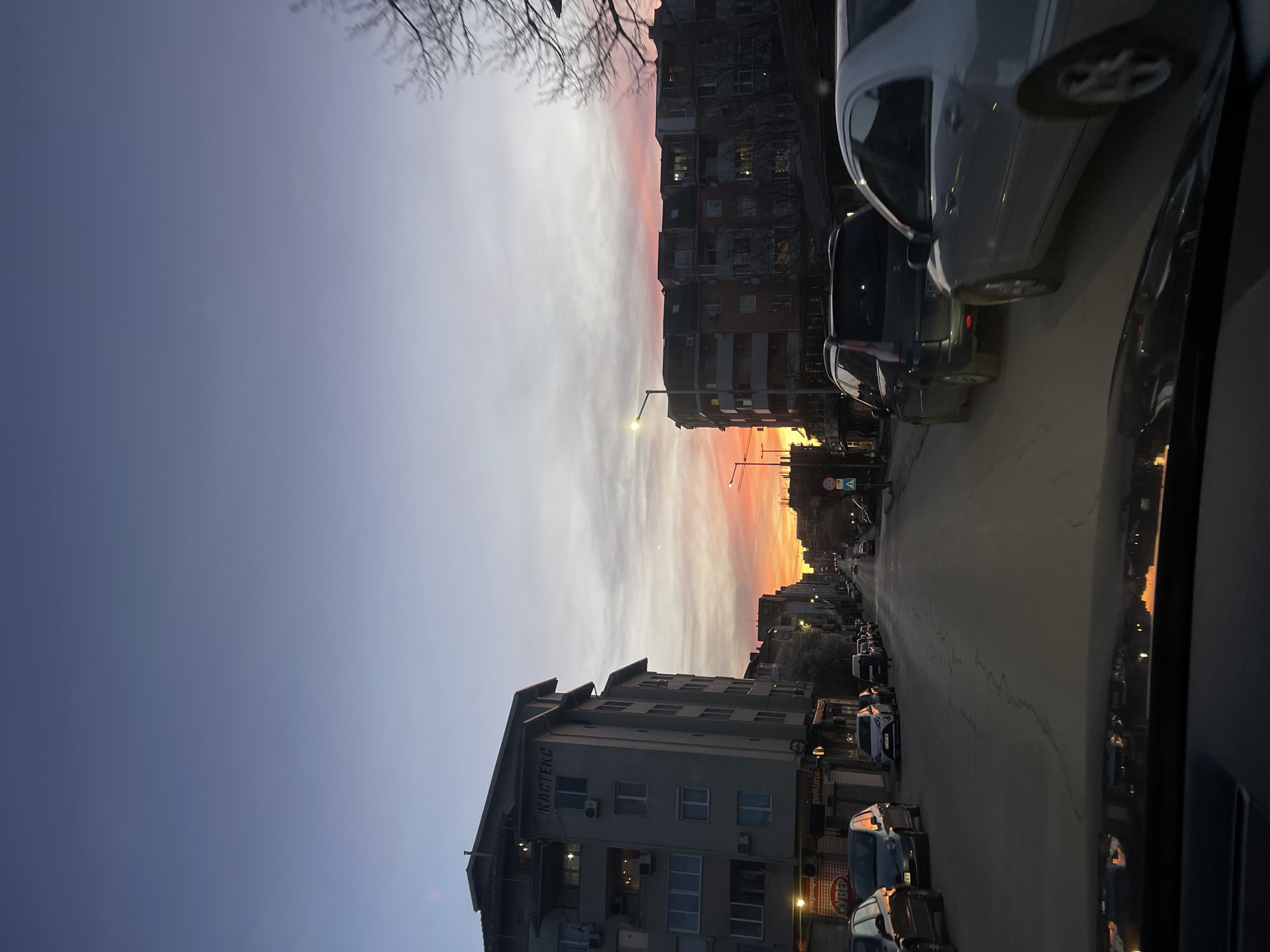 The image depicts a street scene in Veliko Tarnovo, Bulgaria, at dusk. The view is from inside a vehicle, showing the passenger side. The sky transitions from a deep blue to an orange-red sunset in the distance. Buildings line both sides of the street, varying in height and architecture. Cars are parked along the street. The word "КАСТЕКС" is visible on the facade of one building. Streetlights illuminate the scene. Bare tree branches are visible at the top of the frame.