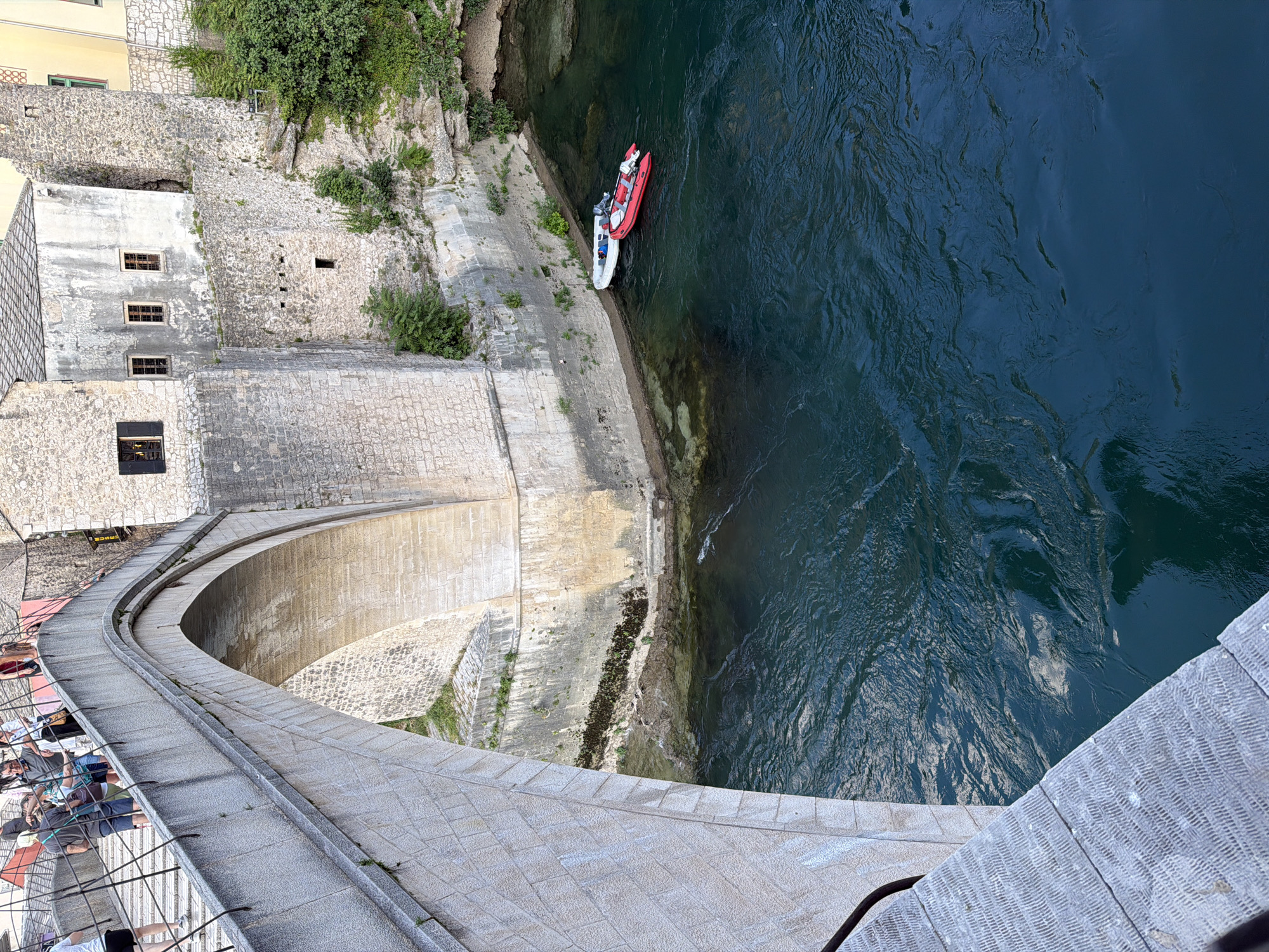 The image shows an elevated view of the Stari Most bridge in Mostar, Bosnia and Herzegovina, with the Neretva River flowing below. The stone bridge arch dominates the left side of the frame, with textured stone blocks and a metal railing visible on the walkway. Several people are walking on the bridge. On the right side, the river is a deep, dark blue, with two inflatable boats moored near the bank. One boat is red, and the other is white. The riverbank is rocky and overgrown with vegetation. The background features ancient stone buildings with arched windows and tiled roofs, characteristic of Mostar's historic architecture. The overall scene depicts a sunny day with clear water and a bustling atmosphere on the bridge.
