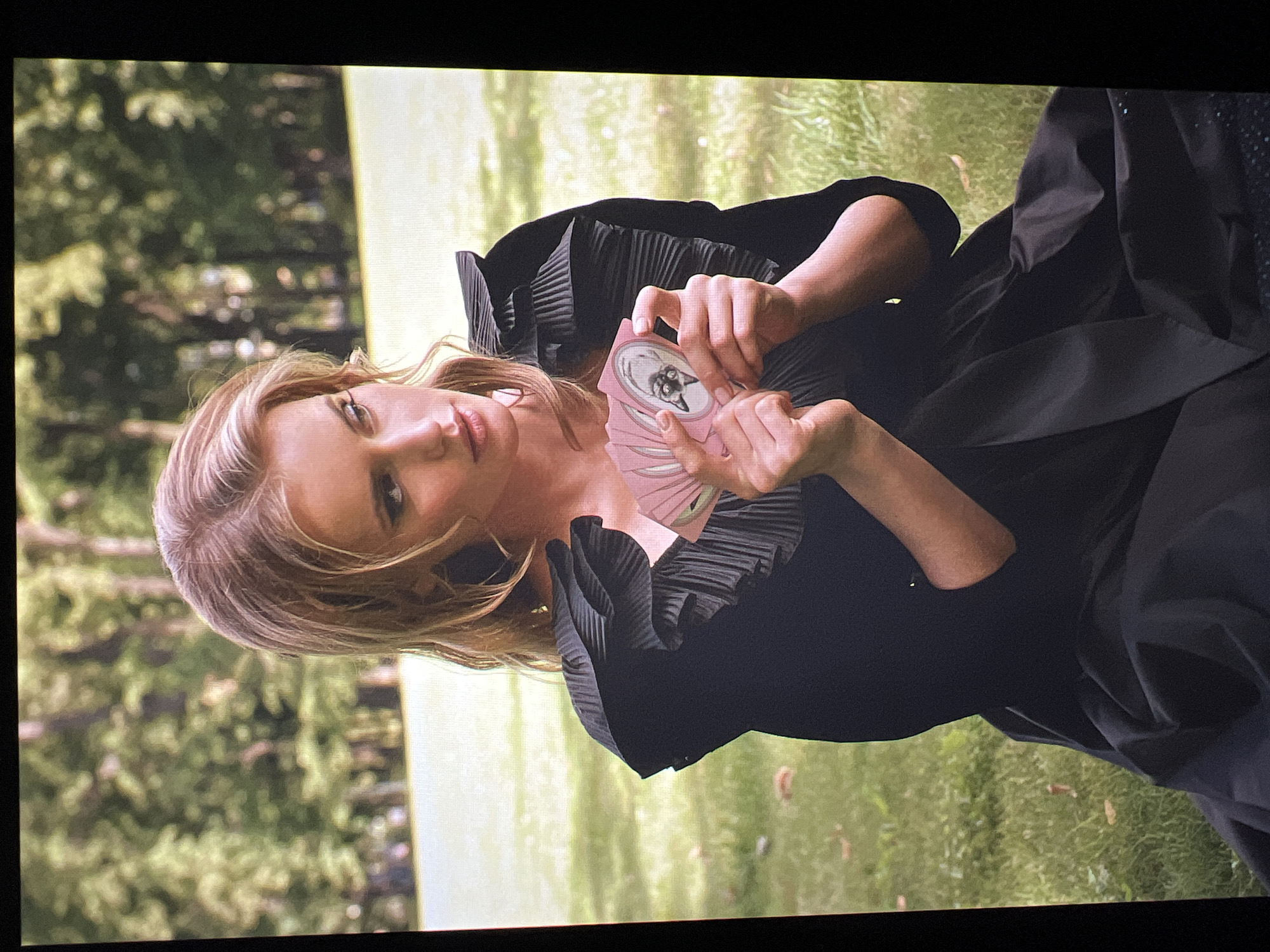 The image shows a young woman with blonde hair sitting on grass in a park, holding a fan of pink cards. The woman is wearing a black dress with ruffled sleeves. The background consists of trees and green grass. The scene appears to be a publicity photograph, possibly for a fashion magazine or brand. The specific location in Saint Petersburg, Russia is not determinable from the image alone.