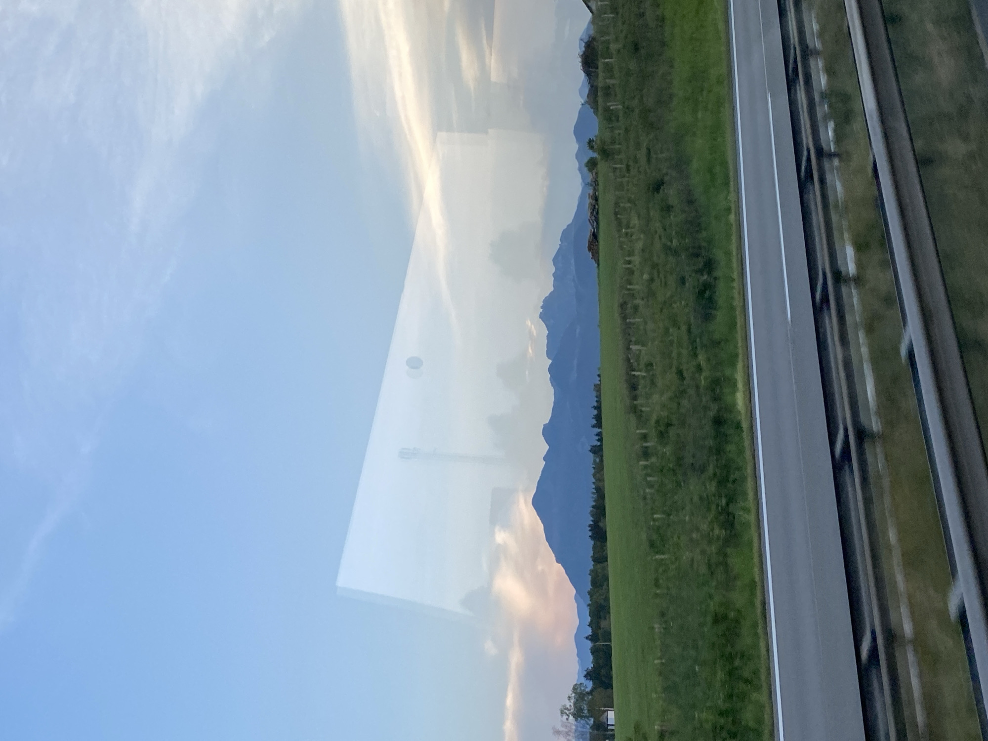 This image captures a view from a vehicle traveling on a highway in Grabenstätt, Germany. The scene is dominated by a reflective window, which overlays the natural landscape with a distinct rectangular reflection.
The foreground features the highway itself, with clear lane markings and a metal guardrail. Beyond the road, a lush green field stretches out, dotted with a simple wooden fence. In the mid-ground, a range of blue-hued mountains forms a dramatic backdrop. The sky above is a gradient of pale blue with wispy white clouds, tinged with soft pink and orange hues, suggesting either dawn or dusk.
The reflection on the window is a significant element, showing a stark, almost geometric structure that appears to be a cell tower. This artificial element contrasts sharply with the natural beauty of the mountains and fields. Additionally, the reflection also captures a segment of the sky and some indistinct shapes, adding a layer of complexity to the visual composition. There are no people or animals visible in the image.