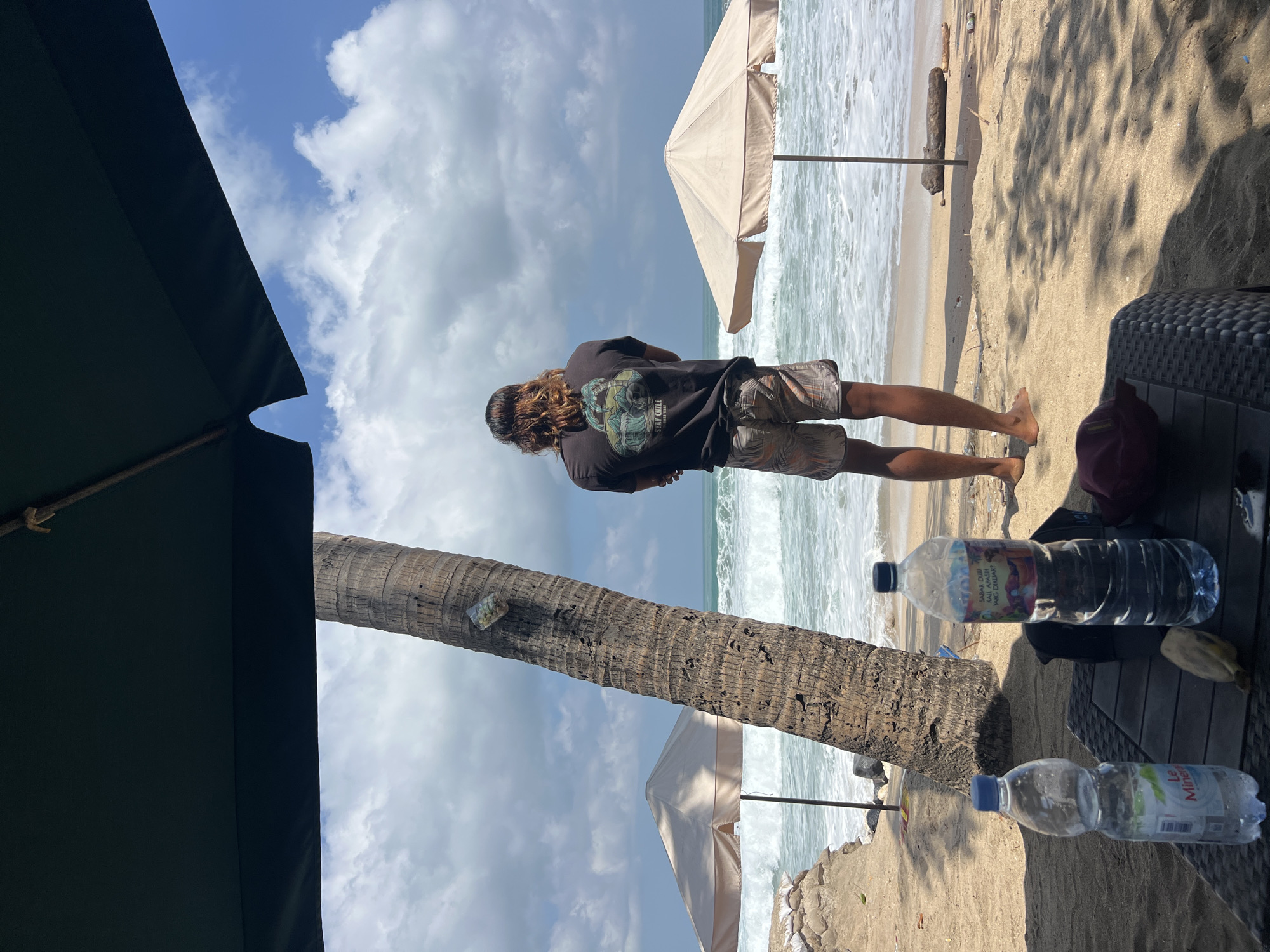 A person with long, brown hair stands with their back to the camera on a sandy beach, looking out at the ocean. The person is wearing a dark t-shirt with a graphic design and patterned shorts. In the foreground, a dark wicker table holds two plastic water bottles and a maroon cap. A palm tree trunk is to the left of the person, and two beach umbrellas are visible further down the beach. The ocean has moderate waves, and the sky is partly cloudy. The scene appears to be a casual outdoor setting, likely in Kuta, Indonesia, given the environment.