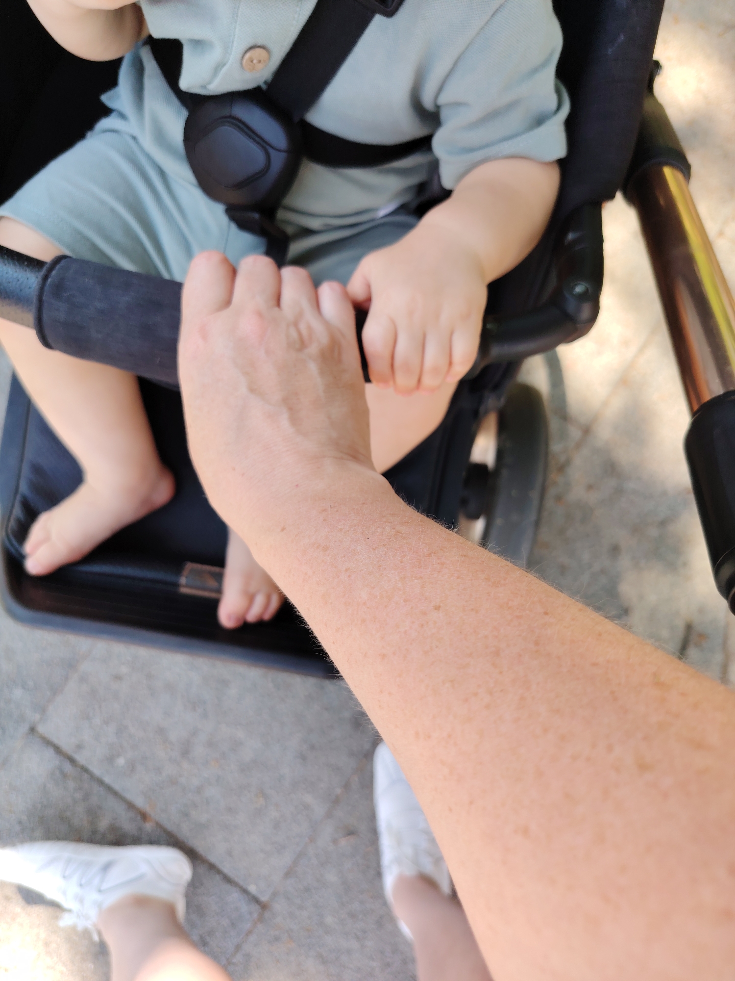 The image shows a close-up view of a baby in a stroller, being held by an adult's hands. The baby is wearing a light green outfit and is secured by a black harness. The adult's hands are visible gripping the stroller's handle. The background consists of a paved walkway with some sunlight filtering through. The scene appears to be outdoors, possibly in a park or on a street in Dolgoprudny, Russia, given the context provided.