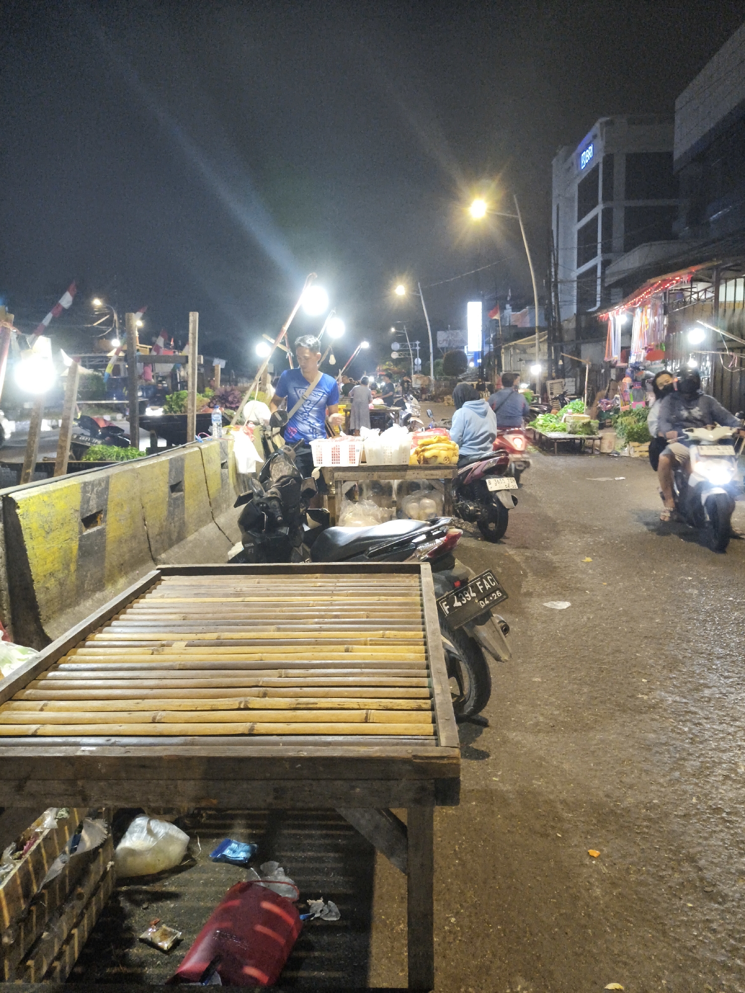 The image depicts an outdoor market scene at night in South Jakarta, Indonesia. A man in a blue shirt stands behind a wooden stall filled with white baskets. Motorbikes are parked along the street, and two people are riding one of them, illuminated by its headlight. The market appears to be set up along a street with buildings visible in the background. There are stalls with bamboo-like structures and various goods displayed, possibly food items. The lighting suggests artificial illumination from streetlights and stall lights. Trash is visible under one of the stalls.