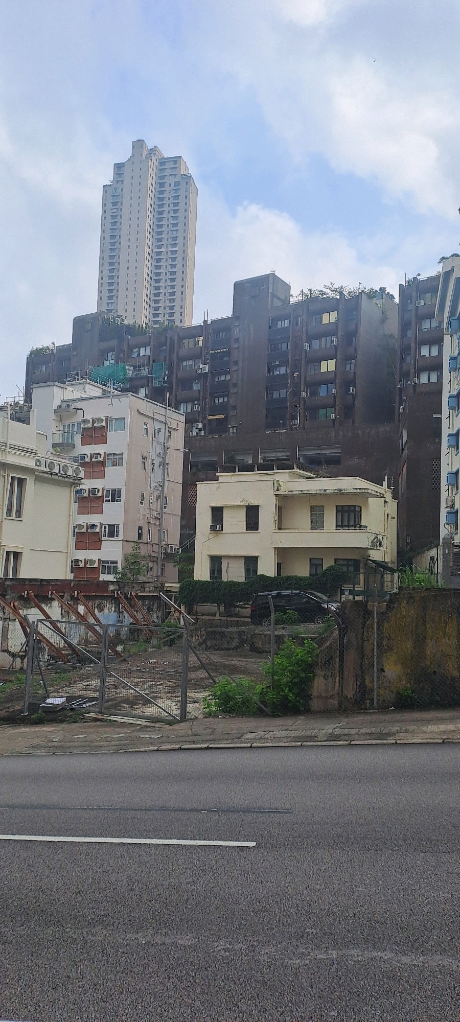 The image depicts a street scene in Hong Kong, China, characterized by a mix of older and newer architecture under a cloudy sky. In the background, a prominent, tall, light-colored skyscraper with numerous windows dominates the skyline, indicating a densely built urban environment. To its left and right are darker, multi-story apartment buildings with balconies and visible air conditioning units. In the foreground, a vacant lot enclosed by a wire fence and a weathered concrete wall suggests ongoing construction or demolition. A black SUV is parked on the lot. The street in the immediate foreground is paved asphalt with visible white lane markings. The overall impression is one of urban development and density typical of Hong Kong. There are no people visible in the frame.