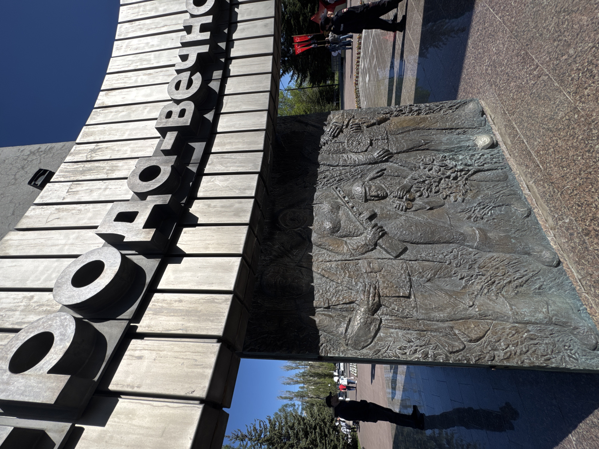 The image depicts a monument, likely a war memorial, in the city of Unknown, Unknown. The monument features large Cyrillic letters stacked vertically, made of concrete or stone, on the left side. To the right, a bronze bas-relief sculpture depicts figures resembling soldiers or historical figures. The ground is paved with granite tiles. People are visible in the background, including a person in a dark uniform and a red flag. The sky is blue.