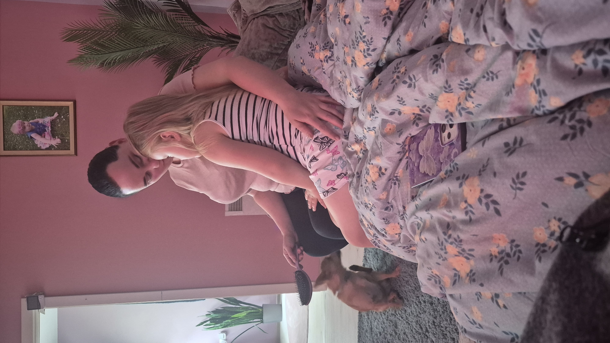 In a bedroom with pale pink walls, a woman with dark hair is seated on a bed, holding a small tan dog that is playfully reaching for a hairbrush held by the woman. Beside the woman is a young girl with blonde hair, wearing a striped tank top and shorts adorned with butterflies. The girl is leaning into the woman, and the woman has her arm around the girl. The bed is covered with a duvet featuring a floral pattern in shades of purple and yellow. A framed picture of a baby is hanging on the wall above them. A potted palm plant is visible to the right of the frame. To the left, a large mirror reflects a doorway and a potted plant with long, green leaves. This scene is likely within a private residence in Švenčionys, Lithuania.