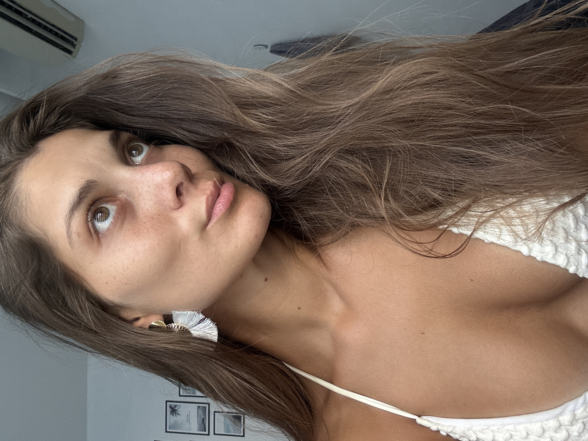 A woman with long brown hair is pictured from a low angle, gazing upwards and to the left. She has fair skin, freckles, and brown eyes. She is wearing a white top with a thin strap. In her left ear, there's a gold earring with white tassels. Behind her, there is an air conditioning unit mounted on the wall and a set of framed pictures. The location is unidentifiable as being in Đà Nẵng, Vietnam based on the provided image.