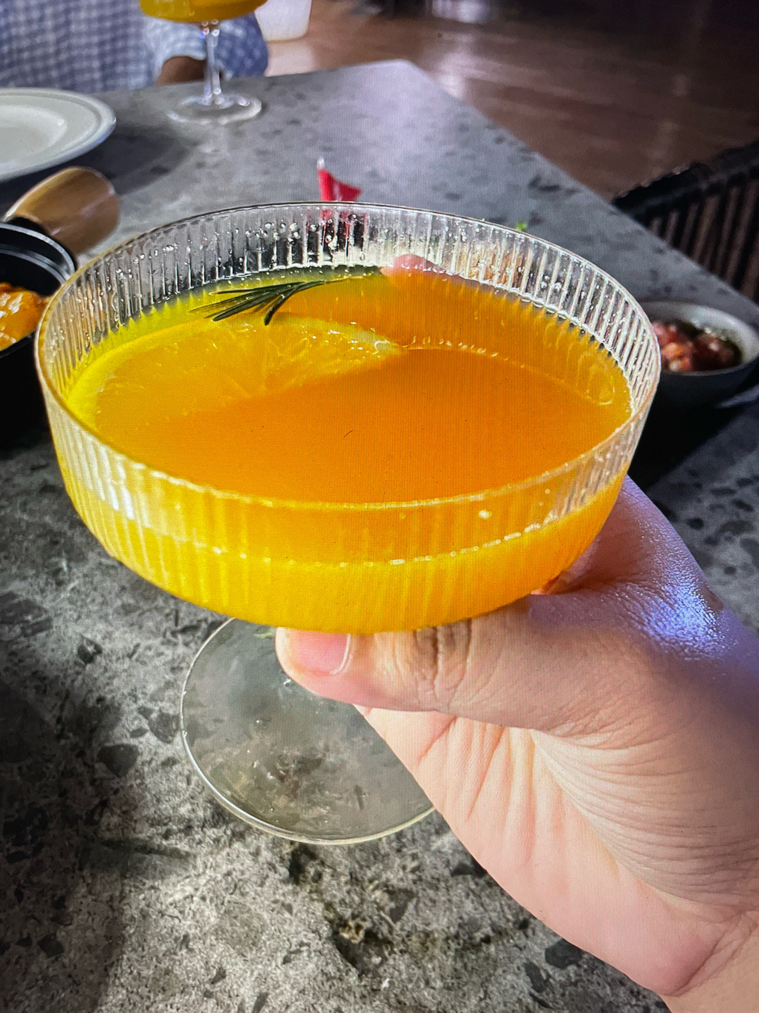 A hand holds a fluted glass coupe filled with an orange liquid, garnished with a sprig of rosemary. The background shows a patterned shirt, a plate, and a small bowl containing a red mixture. The setting appears to be a restaurant or bar with a textured countertop. There is no indication of the city of Gankar, India, in the image.