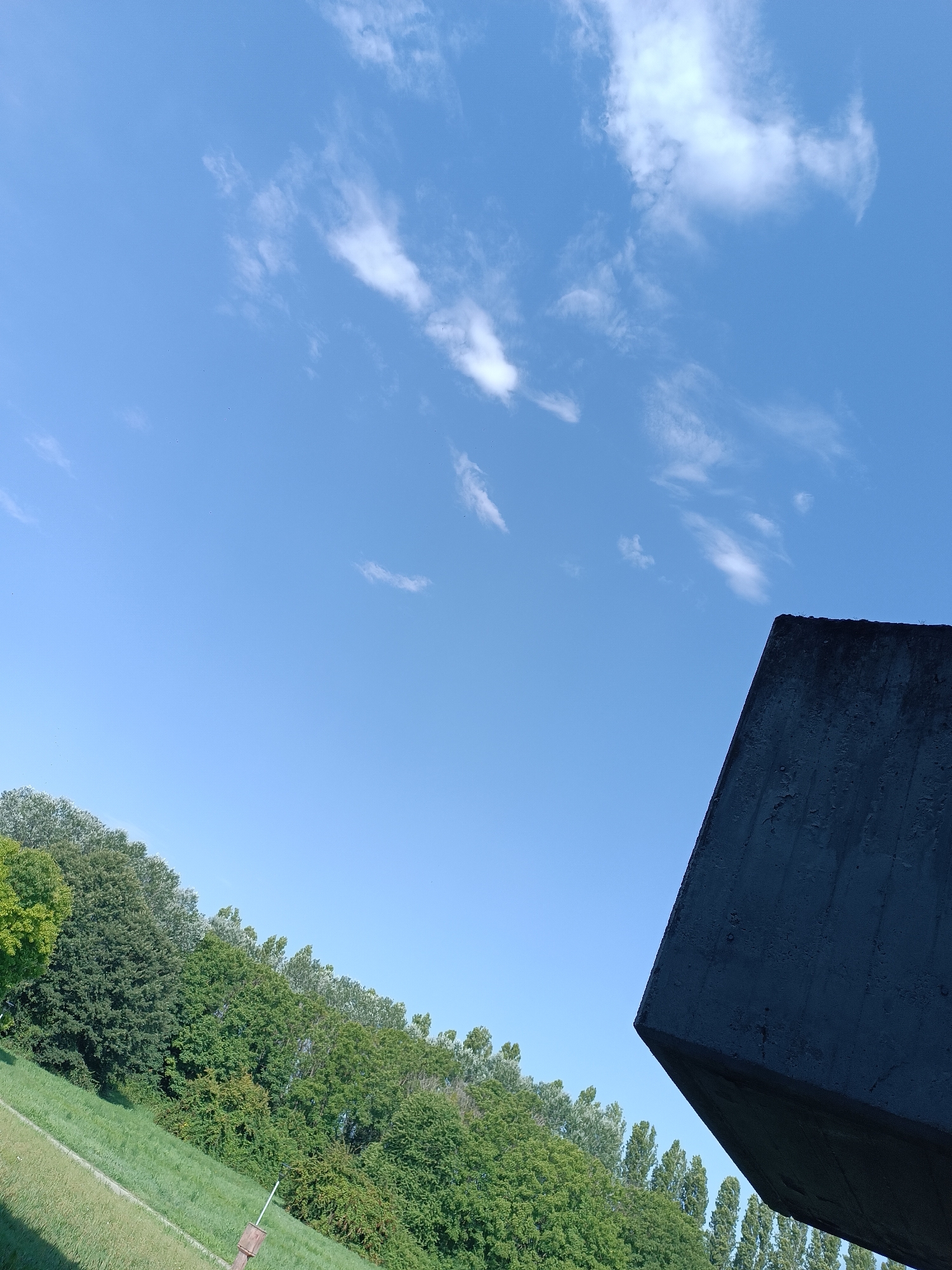 The image displays a clear blue sky with scattered wispy white clouds. In the foreground, a dark, angular concrete structure juts into the frame from the right. Below this structure and extending across the lower portion of the image is a lush green landscape. This landscape consists of a grassy slope interspersed with dense green trees. A white pole with a small brown object at its base is visible on the grassy slope. The trees form a line along the horizon, suggesting a park or natural area. The overall impression is of an outdoor scene on a bright day, likely in a park or open space within Casalmaggiore, Italy, given the reference to the location. No people are visible in the image.