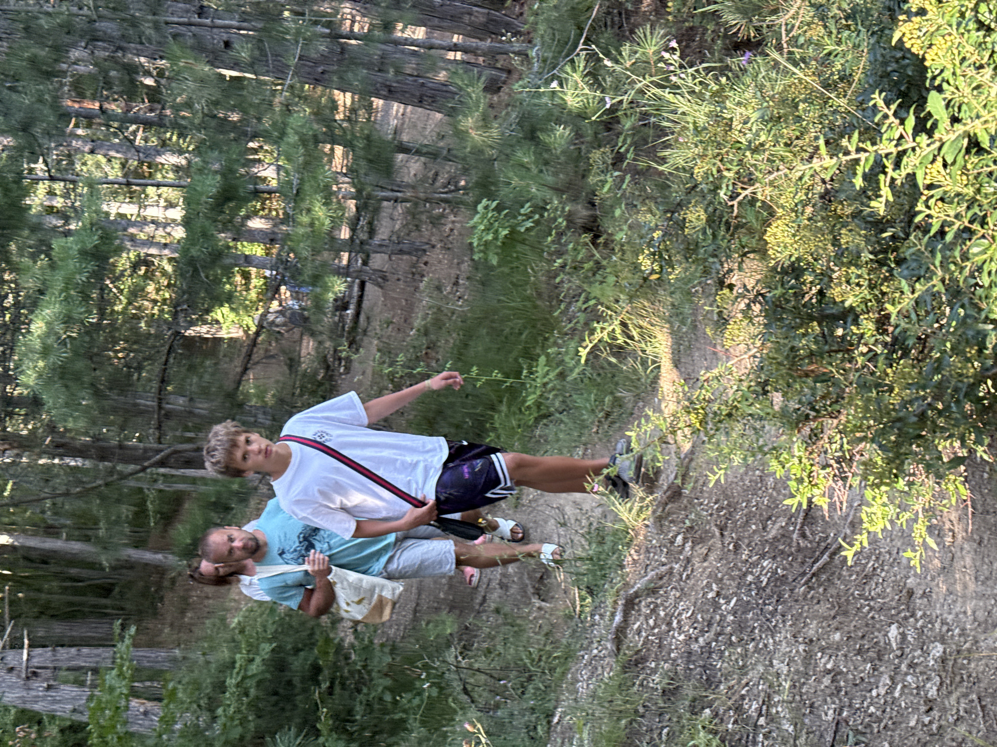 In Zaprudnoe, Ukraine, three individuals are walking along a dirt path in a forest. The person in the foreground is a young male with light brown hair, wearing a white t-shirt and dark shorts. He is carrying a strap across his body. Behind him, a man in a blue t-shirt is walking, and a woman in a white top is visible further back. They are all in a wooded area with tall trees and dense green foliage. The path appears to be inclined.