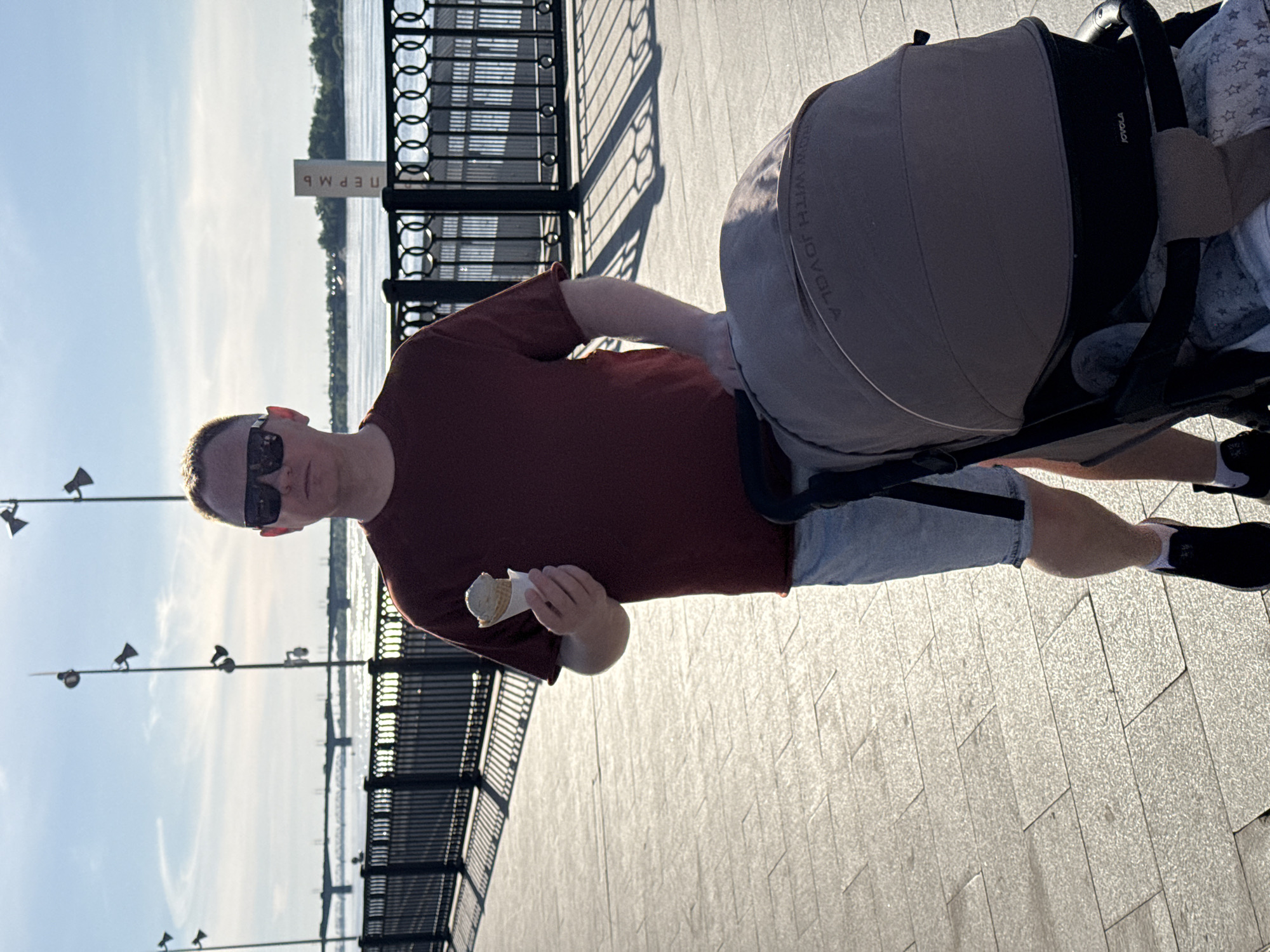 A man stands on a paved walkway in Perm, Russia, pushing a baby stroller. He wears sunglasses, a maroon t-shirt, and gray shorts. He holds an ice cream cone in his left hand. A black metal fence separates the walkway from a body of water. Streetlights line the walkway. A sign with the Russian word "Пермь" (Perm) is visible. The stroller is to the right of the man. The lighting suggests it is daytime.