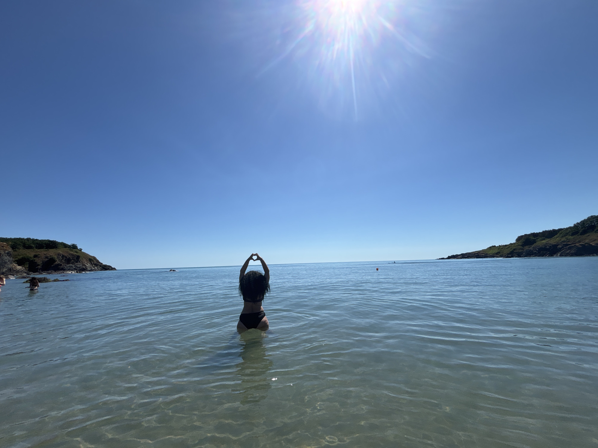 A wide-angle, full shot captures a person standing in shallow, clear water at a beach in Rezovo, Bulgaria, on a bright, sunny day. The sun is high in a clear blue sky, creating a bright lens flare at the top of the frame. The person, viewed from behind, is wearing a black bikini and has long, dark, curly hair. They are standing facing away from the camera, with their arms raised above their head and hands forming a heart shape. The water is calm and reflects the sunlight. In the distance, two rocky, tree-covered headlands flank the bay, and a small orange buoy is visible in the water. The seabed is visible through the clear water.