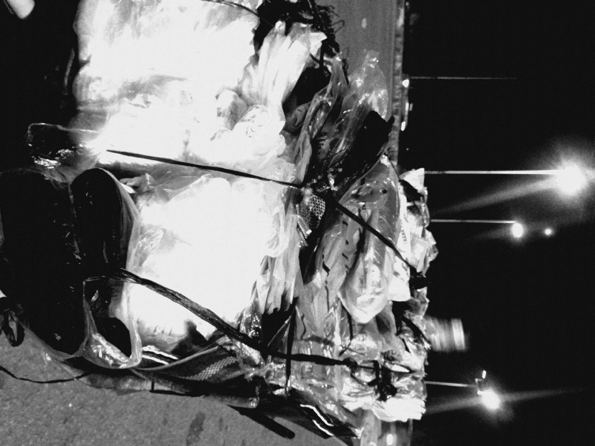 This is a grainy, black and white photograph depicting a large pile of discarded materials, primarily plastic bags, at night in Bukuru, Nigeria. The image appears to be rotated, showing what would normally be the bottom of the pile on the left side.
The main subject is a substantial mound of refuse, dominated by a tangled mass of white and translucent plastic bags that appear somewhat illuminated, possibly reflecting light or holding lighter contents. Interspersed within the plastic are darker objects, which could be shoes, fabrics, or other solid debris. Thin, dark, rod-like structures also protrude from and weave through the pile. The texture of the plastic is evident, appearing crinkled and voluminous.
The pile rests on what appears to be a paved ground or road surface, visible along the bottom edge of the image (when correctly oriented). The background to the right is largely dark, with several distinct, bright points of light visible in the distance. These are likely streetlights, casting prominent, narrow beams of light horizontally across the dark expanse, indicating the scene takes place at night.
There is no discernible activity taking place; the image captures a static scene of accumulated waste. The overall impression is one of an urban or semi-urban environment, highlighting issues of waste management. Faintly visible on a light-colored plastic bag within the pile, the words "THANK YOU" can be discerned.