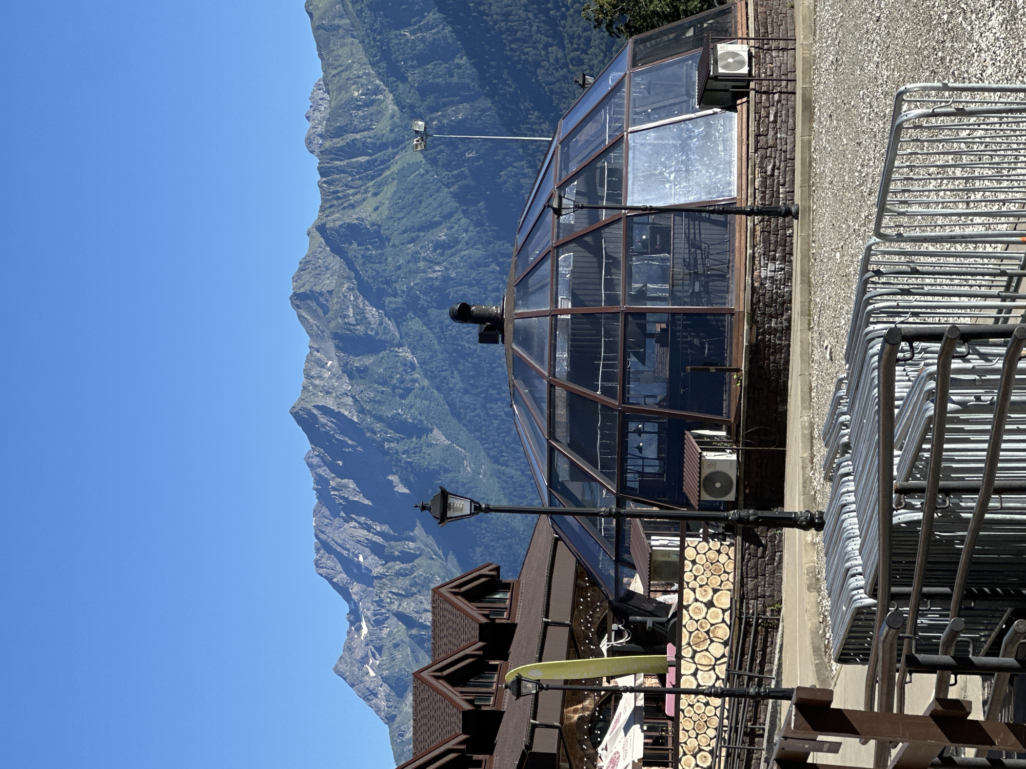 The image shows a sunny day in Estosadok, Russia. In the foreground, there are several metal barriers, likely for crowd control, stacked on a gravel surface. Behind the barriers, a building with a distinctive geodesic dome made of glass and dark metal framing is visible. The dome appears to be a restaurant or cafe, with tables and chairs visible inside. To the left of the dome is a two-story building with a brown, shingled roof and wooden accents on its facade. There are also street lamps and outdoor seating areas visible. In the background, a majestic mountain range with green slopes and snow-capped peaks rises under a clear blue sky. The overall scene suggests a resort or tourist area in a mountainous region during pleasant weather.