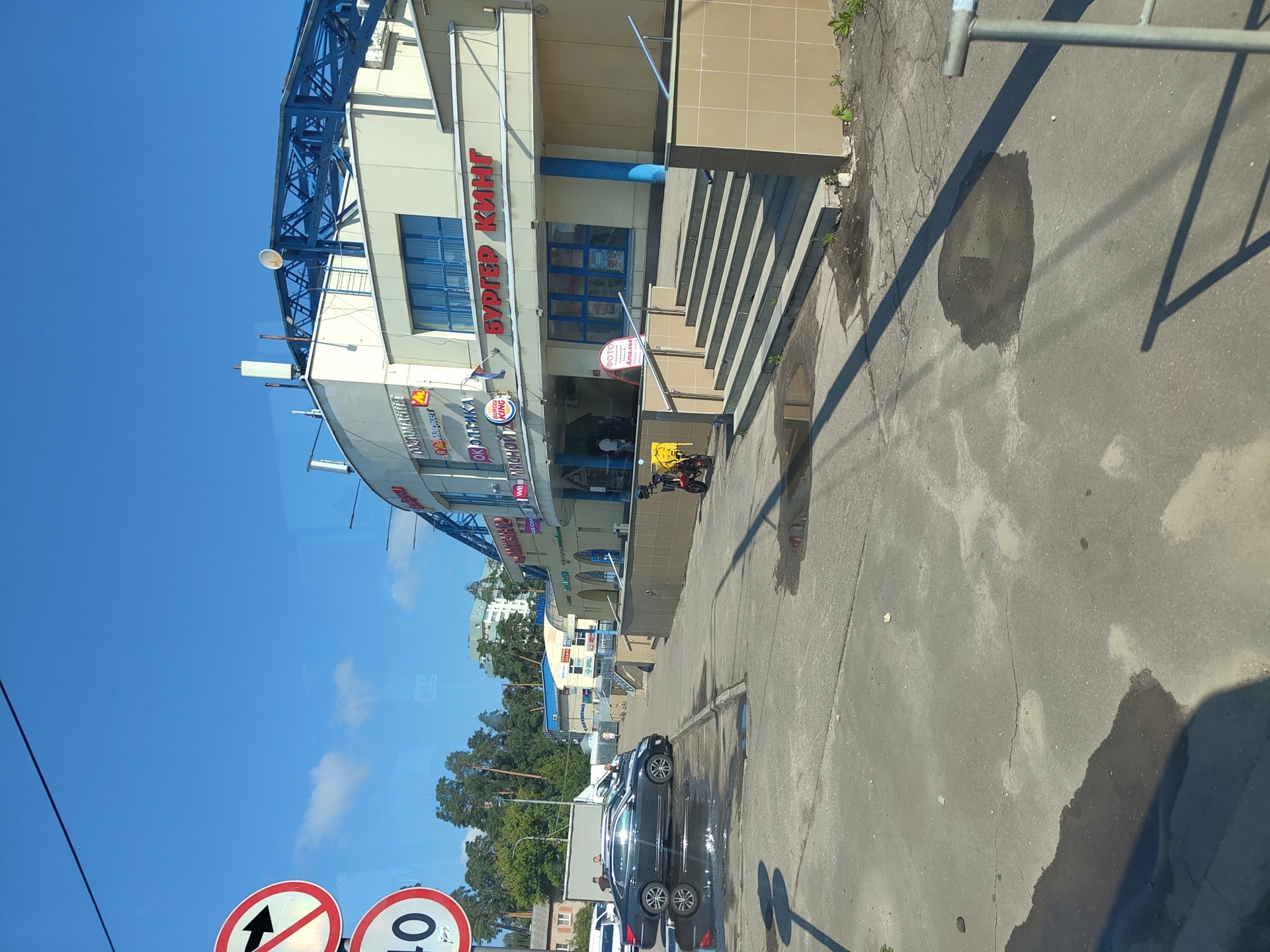 This is a photograph of a commercial building in Zhukovsky, Russia, on a clear day with a blue sky and some clouds. The building appears to be a shopping center or market, with various storefronts visible. One prominent sign reads "БУРГЕР КИНГ" (Burger King). Other signs include "ЗОМЯН" (presumably a store name) and "ОК ОПТИКА" (OK Optics). The building has a light-colored facade and a blue, modern-looking roof structure.
In the foreground, there is a paved area with several puddles on the ground, suggesting recent rain or cleaning. A dark-colored sedan is parked on the left side of the image. To the right of the building's entrance, there are a set of stairs leading up to the building. A red scooter with a yellow delivery box is parked near the bottom of the stairs. Two people are walking away from the camera on the sidewalk in front of the building.
In the upper left corner, two traffic signs are visible. One is a circular red and white sign with a black arrow pointing right, indicating a prohibition. Below it is a circular sign with the number "40" in black on a white background, likely indicating a speed limit. In the background, trees and other buildings are visible, including a taller, light-colored building in the distance. The overall scene depicts an outdoor urban environment.