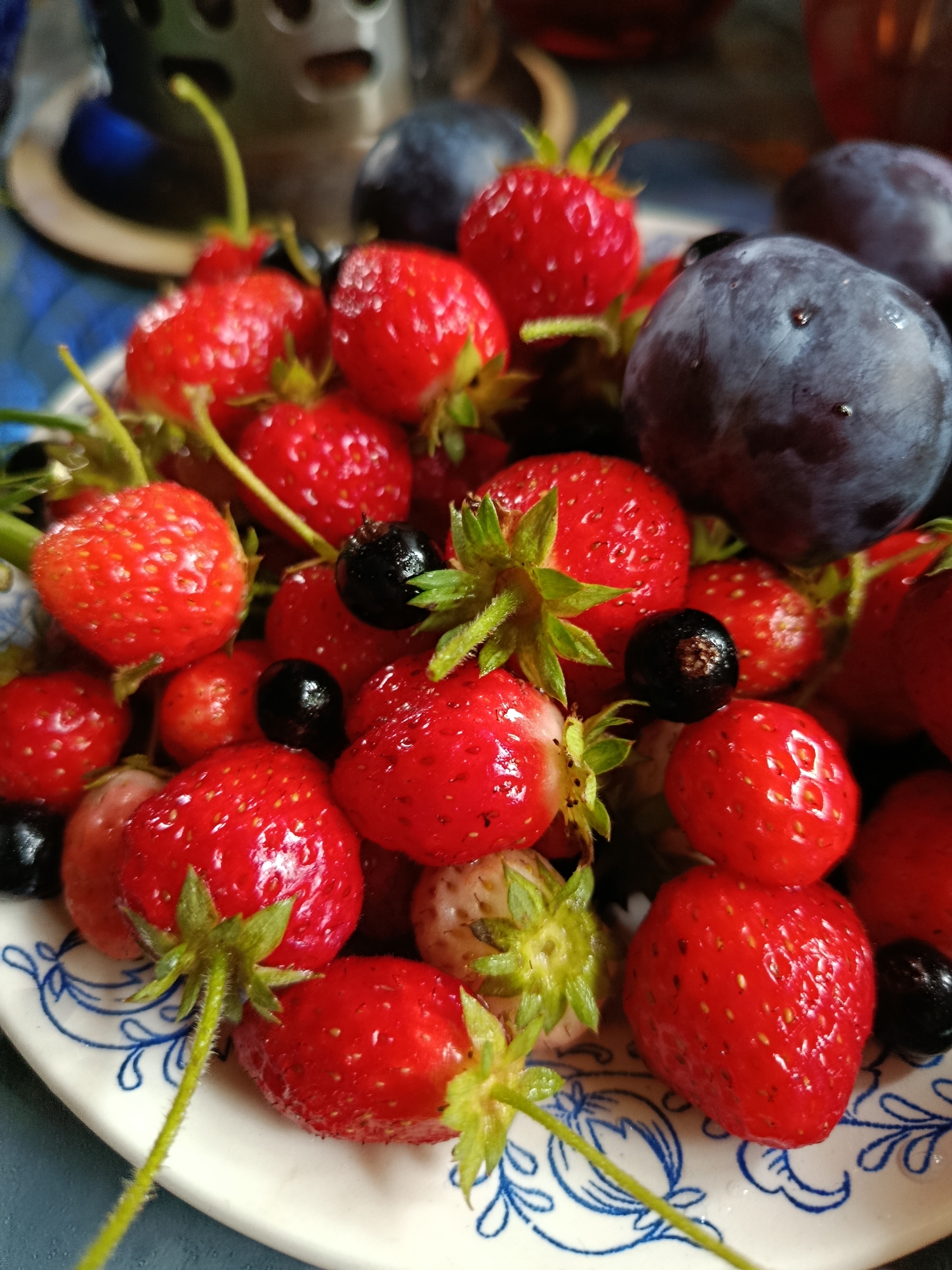 A close-up, overhead shot showcases a plate brimming with fresh strawberries and dark plums, interspersed with blackcurrants. The fruits are glistening, suggesting they have been recently washed. The plate itself is white with a delicate blue floral pattern along the rim. In the background, out of focus, are hints of a blue surface and a metallic object, possibly a utensil holder. The overall impression is one of a rustic, homemade presentation of summer fruits.
