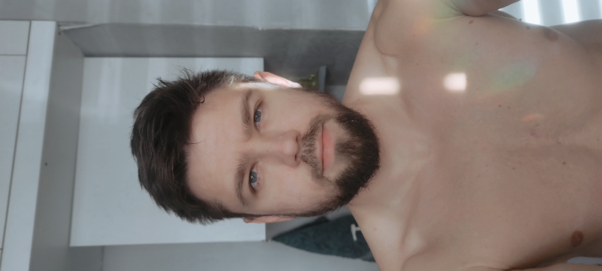 A fair-skinned man with short dark hair and a beard is shown from the chest up, facing the camera. He has blue eyes. He is shirtless. The background includes a white tiled wall, a gray wall, and a green towel. The location is not discernible as Fryazino, Russia based on the image.