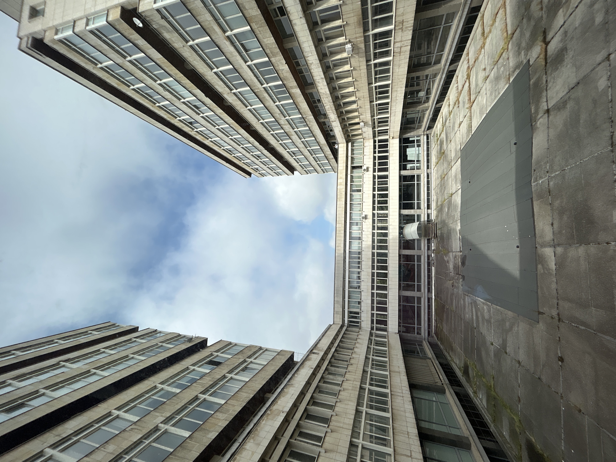 The image presents an upward-looking architectural view captured between two tall, multi-story buildings in Sofia, Bulgaria. The perspective emphasizes the towering height of the structures, with their facades converging towards the top.
Both buildings exhibit a utilitarian architectural style, featuring numerous rectangular windows arranged in orderly horizontal bands across their surfaces. The primary material appears to be concrete or stone, ranging in color from lighter beige-grey on the left building to a darker, more textured grey on the right. The right-hand building shows signs of weathering, including moss or discoloration on its concrete panels, and features a large, dark grey rectangular panel or door on one of its lower sections.
Between the buildings, a section of the sky is visible, showing a mix of blue sky and white, fluffy clouds, indicating a daytime setting with partial cloud cover. The extreme low-angle shot creates a sense of scale and depth. No people or specific activities are depicted, and there is no visible text.