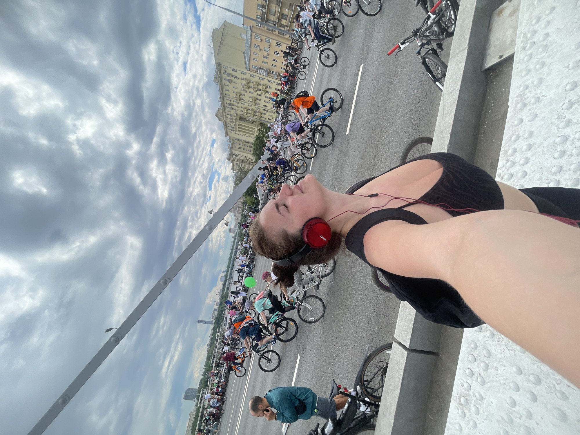 A selfie shows a woman with auburn hair in a ponytail, red headphones, and black workout gear, looking upwards with her mouth slightly open. She stands on a bridge with a low grey concrete barrier visible on the right. Behind her, a mass of cyclists ride on a wide road, and a light-colored building can be seen in the background. The sky above is cloudy. A man wearing a blue shirt stands to the left of the woman, looking at his mobile phone while leaning on his bike. The city of Unknown, Unknown.