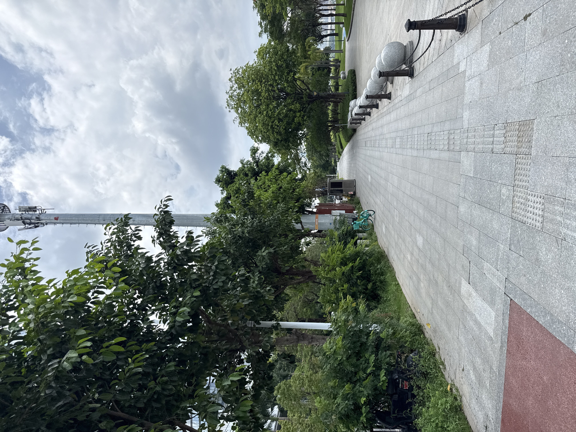 The image depicts a paved pedestrian walkway in Haizhu District, China, bordered by lush green trees and vegetation on the left and a grassy area with more trees on the right. The walkway itself is constructed with large, light gray stone pavers, featuring tactile paving for visually impaired pedestrians. Along the right side of the walkway, a row of decorative, spherical bollards mounted on dark posts are connected by black chains. In the distance, the walkway curves slightly and appears to lead towards a body of water or open space.
On the left side of the walkway, a small, light blue bicycle is parked near some bushes. Behind the bicycle and to the left, a dark-colored scooter is visible, partially obscured by greenery. A tall, grey cellular tower stands prominently against the cloudy sky in the background, to the left of the walkway. Several people are not visible in the image. The environment appears to be a public park or waterfront promenade, with well-maintained landscaping and urban infrastructure. The lighting suggests an overcast day.