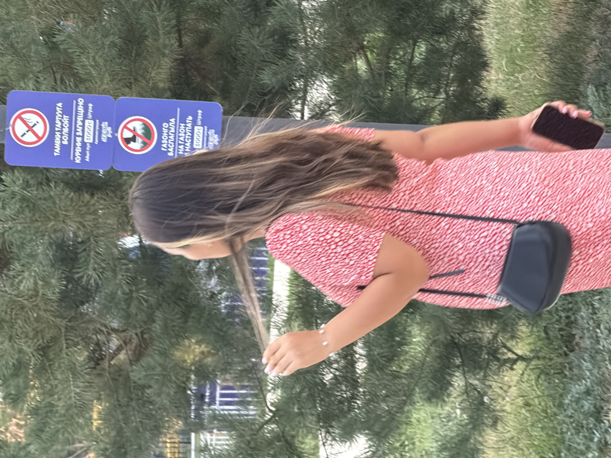 A woman is captured in a medium shot, facing away from the camera and looking to her left. She has long, dark brown hair with lighter highlights and is wearing a red and white patterned dress. A small black purse hangs from a strap across her body. In her right hand, she holds a dark-colored smartphone. Her left hand is raised, and she is touching her hair.
Behind her, partially obscured by the woman and a large evergreen tree, are two blue signs stacked vertically. The top sign displays a "no smoking" symbol and text in what appears to be Cyrillic script. The bottom sign features an icon of a person stepping on grass, also with a "no" symbol, and Cyrillic text below. The background is dominated by the dense green foliage of the tree, with some hints of other vegetation and structures visible further in the distance. The lighting suggests it is daytime.