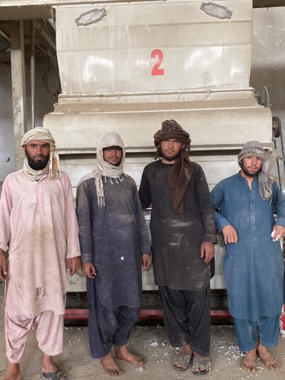 Four men stand barefoot in front of industrial machinery labeled with a red "2." The men wear traditional clothing and head coverings. Dust or white powder coats their clothing and the floor. The scene is located in what is likely Shurandam, Afghanistan.