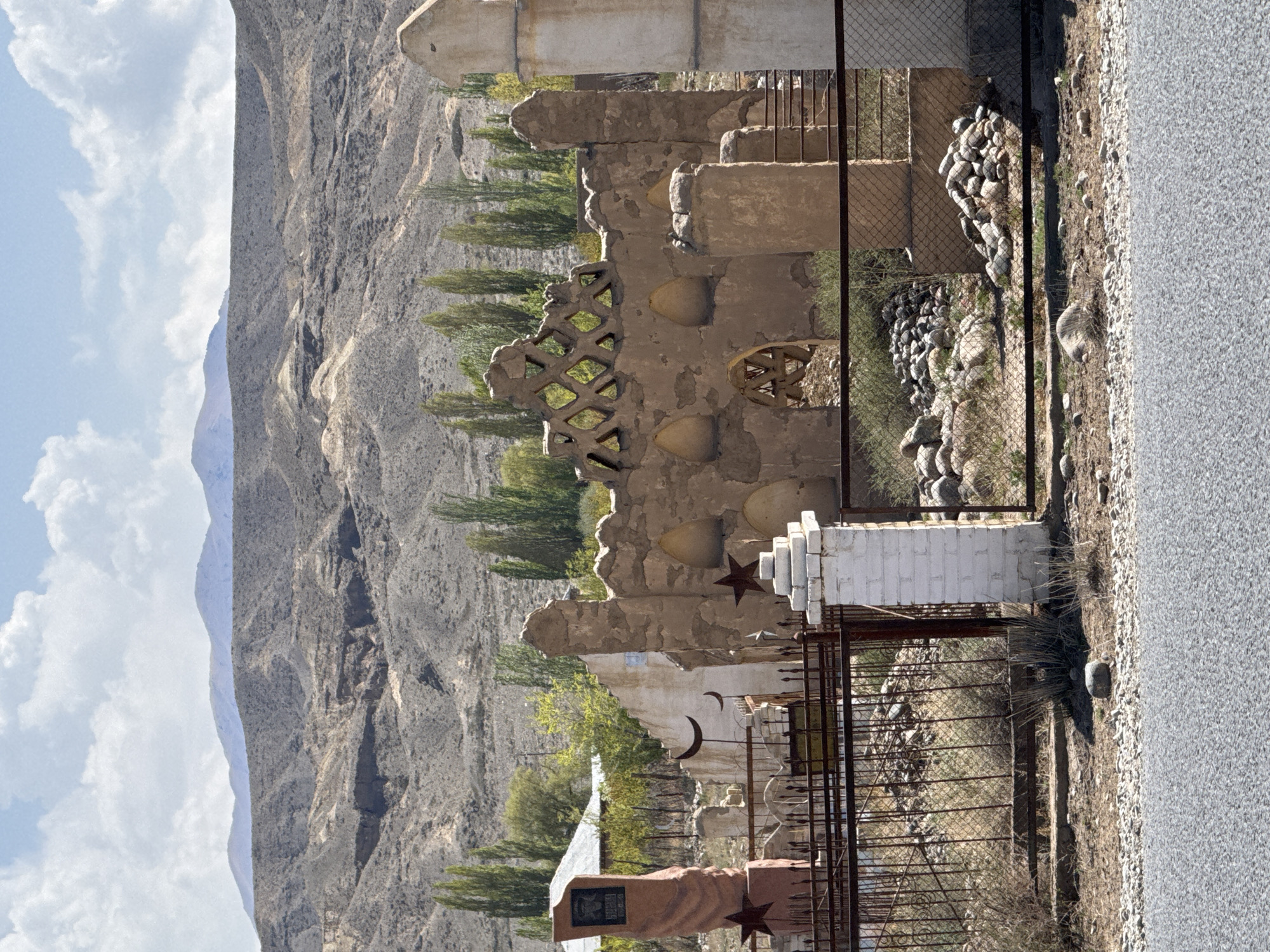 The image depicts a solemn landscape featuring a mountainous backdrop, a paved road, and what appears to be a cemetery or memorial area. The scene is set in Kara-Talaa, Kyrgyzstan, under a partly cloudy daytime sky.
In the foreground, a gray paved road runs horizontally with a gravel shoulder. Beyond the road, a chain-link fence separates the road from a series of structures, which include various grave markers and walls. Many grave markers are simple rectangular stones, while others are more ornate. Prominent symbols visible include five-pointed stars, often on white pedestals or attached to grave markers, and crescent moons, some cut into metal or affixed to structures. One notable reddish-brown grave marker in the bottom-left features a dark plaque with indistinct text, likely in a local script. Another structure is made of white bricks.
A rough, textured wall with decorative cutouts and embedded round or beehive-like shapes forms part of the memorial area. Behind these structures, scattered tall, slender trees with green foliage are visible, adding a touch of life to the otherwise arid surroundings. Further back, towering, barren mountains with deep crevices and varied rock formations dominate the background, their gray and brown hues contrasting with the blue sky and white clouds.
No human activity is depicted in the image, suggesting a quiet and contemplative atmosphere. The symbols on the graves and the rugged terrain are consistent with the Central Asian location.