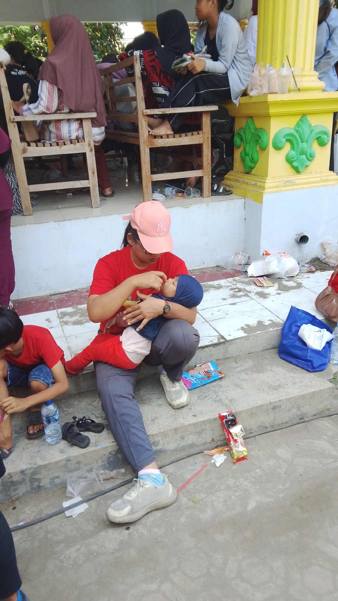 The image captures a scene on an outdoor concrete stairway in Mancar, Indonesia. A woman, wearing a red t-shirt, grey pants, and a pink baseball cap, is seated on the stairs, holding a baby dressed in blue. The woman appears to be feeding or attending to the baby. To the left, a child in a red t-shirt and denim shorts is also seated on the stairs, with a plastic water bottle and a pair of black sandals nearby. In the background, other people are visible seated on wooden benches under a covered area with a yellow pillar. Various objects, including plastic bags, bottles, wrappers, and a blue tote bag, are scattered around the stairs and ground, indicating a public gathering or resting spot. The overall environment suggests an outdoor public space, possibly a resting area or waiting space.
