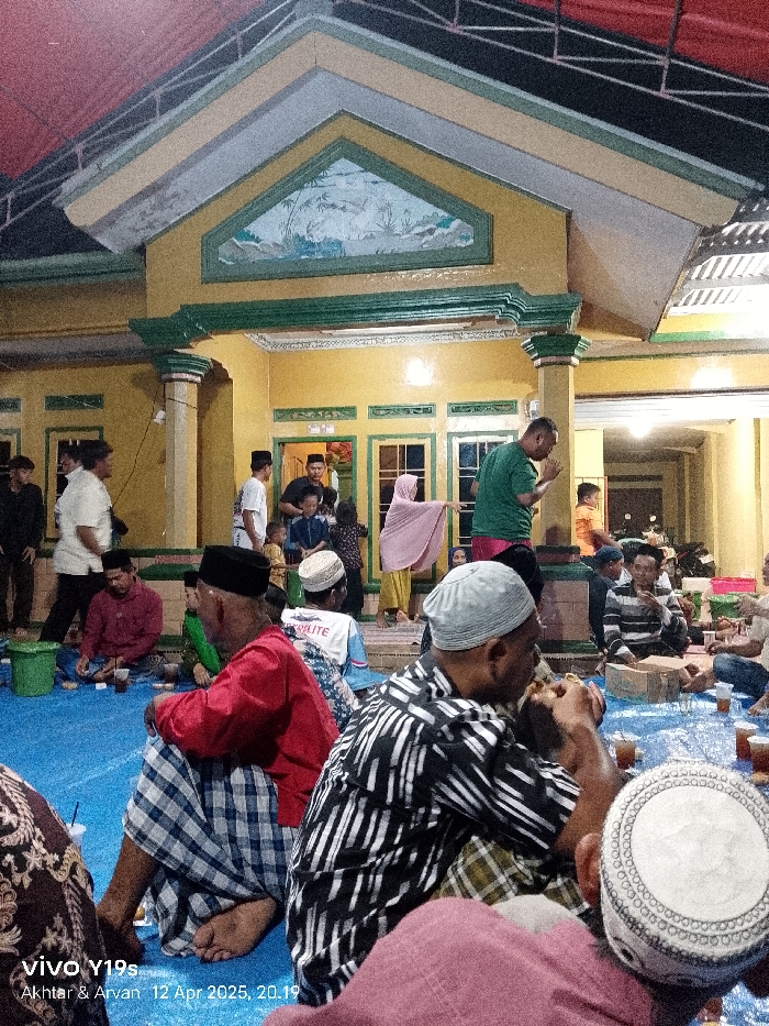 The image shows a gathering of people at a home in Anjatan, Indonesia (latitude: -6.419, longitude: 107.978). The home is a two-story structure with a yellow facade and a green trim. The front porch is covered by a red and black awning. There are 15 people visible in the image. Most of them are men. They are all wearing traditional clothing. They are seated on a blue tarp, drinking and eating. There are several empty glasses and food containers on the tarp. Two men are standing and talking to each other. There is a woman in a white headscarf standing on the porch. The man closest to the camera is wearing a black and white striped shirt. He is eating something with his right hand. The man next to him is wearing a red shirt and plaid pants. He is holding a glass in his left hand. The other people in the image are mostly obscured by the men in the foreground. There is a white, plastic bucket in the foreground. There is a sign on the wall of the home that reads "Viva Y19s". There is another sign on the tarp that says "Akhtar & Arvan 12 Apr 2025, 20:19". The image is taken at night, but the lights are on inside the home and on the porch. The air is clear.