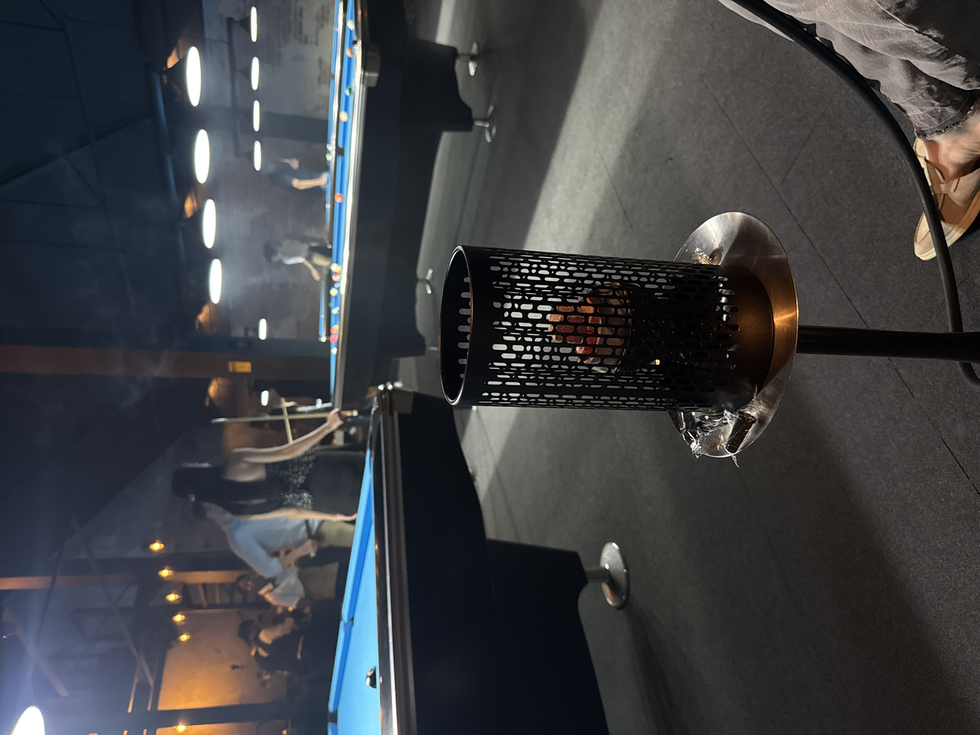 The image captures an indoor scene, likely a pool hall or billiards room, during the evening or night. In the immediate foreground, a striking black cylindrical object with a perforated pattern stands on a dark pole, possibly a decorative lamp, a charcoal holder for a hookah, or a small heater. It emanates a warm, orange glow from within, visible through the perforations, and rests on a brass-colored base. Crumpled clear material, resembling plastic, is gathered around the base of this object.
To the right of this foreground element, the lower part of a person's leg and foot are visible, clad in grey trousers and a light-colored slip-on shoe, suggesting someone is seated or standing nearby.
The background reveals a spacious area with multiple pool tables, distinguished by their vibrant blue felt. Several blurred figures of people are gathered around these tables, engaged in playing billiards or watching. Bright, oval-shaped ceiling lights cast a strong illumination over the pool tables, creating reflections on their surfaces. Further back, additional, warmer lights can be seen. A light haze or smoke is present in the air, particularly noticeable in the upper left of the frame. The floor is dark and appears to be tiled or carpeted. No explicit text is visible in the image. The overall atmosphere appears social and relaxed.
