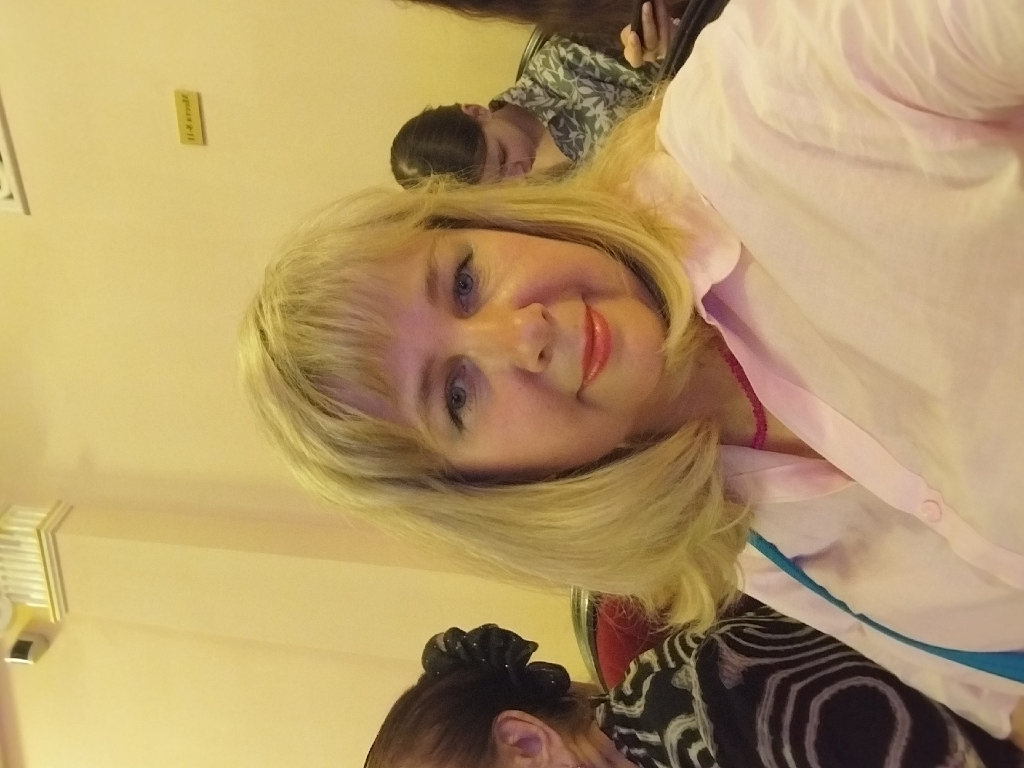 The image shows a close-up, rotated portrait of a blonde woman with blue eyes and red lipstick. She is wearing a light pink shirt. Behind her, a person with dark hair wearing a patterned shirt is visible, and another person with dark hair is seen resting their head on their hands. The setting appears to be indoors, with a pale yellow wall and a vent on the left side of the frame. A small, rectangular object with text is affixed to the wall. The overall lighting suggests an indoor setting, possibly a meeting room or conference. The woman's expression is a smile, suggesting a positive and pleasant mood. The text on the wall is difficult to read clearly but appears to be "11-8 370027" or similar characters. There are no clear indicators of time of day or weather conditions. The context suggests a social gathering or event.