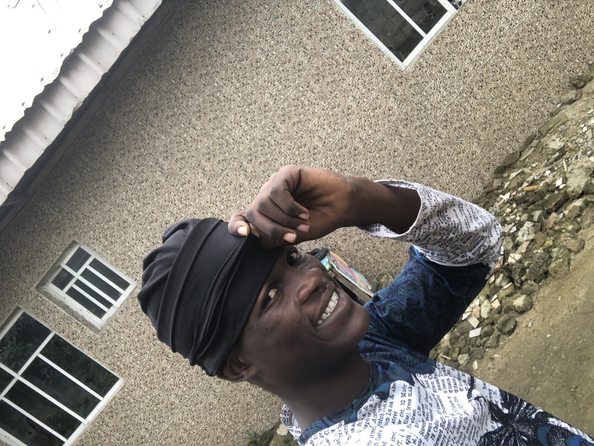 An outdoor, low-angle, medium shot shows a Black man wearing a black headwrap and a white and blue patterned shirt. He is smiling and looking at the camera. The man is touching the brim of his headwrap with his right hand. The background is a textured stone wall with two windows and a corrugated metal roofline. The ground in the foreground consists of loose rocks and dirt. The scene appears to be in Ado, Nigeria.