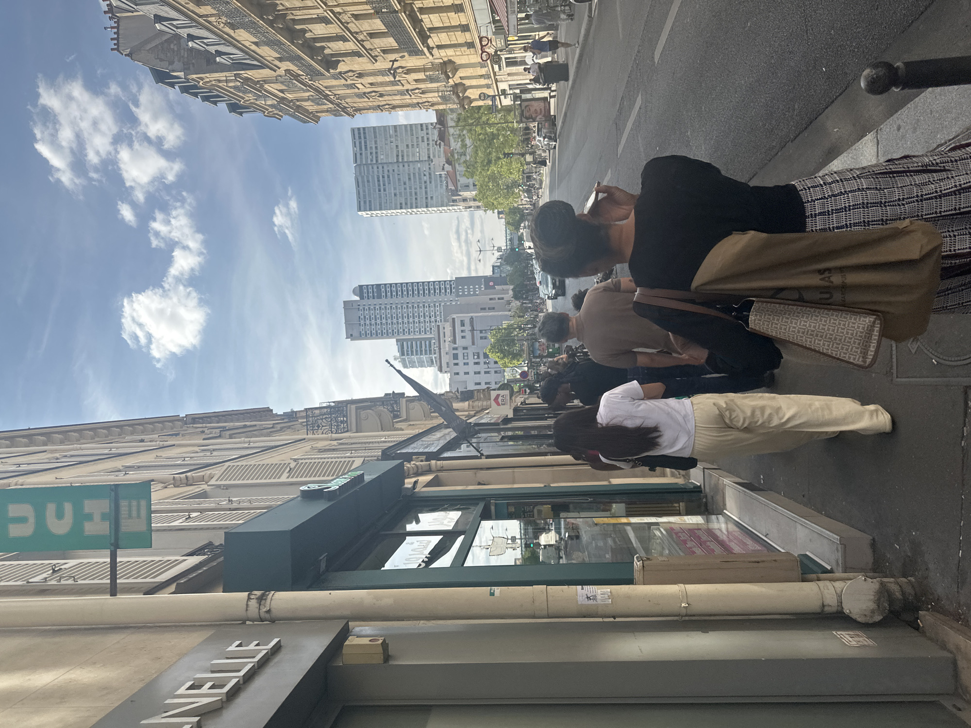 In Paris, France, a group of people are walking along a sidewalk next to a street. The buildings on the right side of the street are tall and ornate, typical of Parisian architecture, with balconies and mansard roofs. The buildings on the left appear to be commercial establishments with storefronts. In the foreground, a woman in a white t-shirt and loose beige pants walks away from the camera, carrying a black purse. Behind her, another woman with dark hair, wearing a black top and a plaid skirt, walks away from the camera, holding a mobile phone to her ear and carrying two tote bags. Further back, a man in a brown t-shirt and dark pants also walks away from the camera. In the background, tall, modern high-rise buildings can be seen, suggesting a mix of historic and contemporary architecture within the city. The sky is blue with scattered white clouds. The street is paved and there are cars and traffic lights visible. A green sign with the word "UCHE" in white letters hangs vertically from one of the buildings.