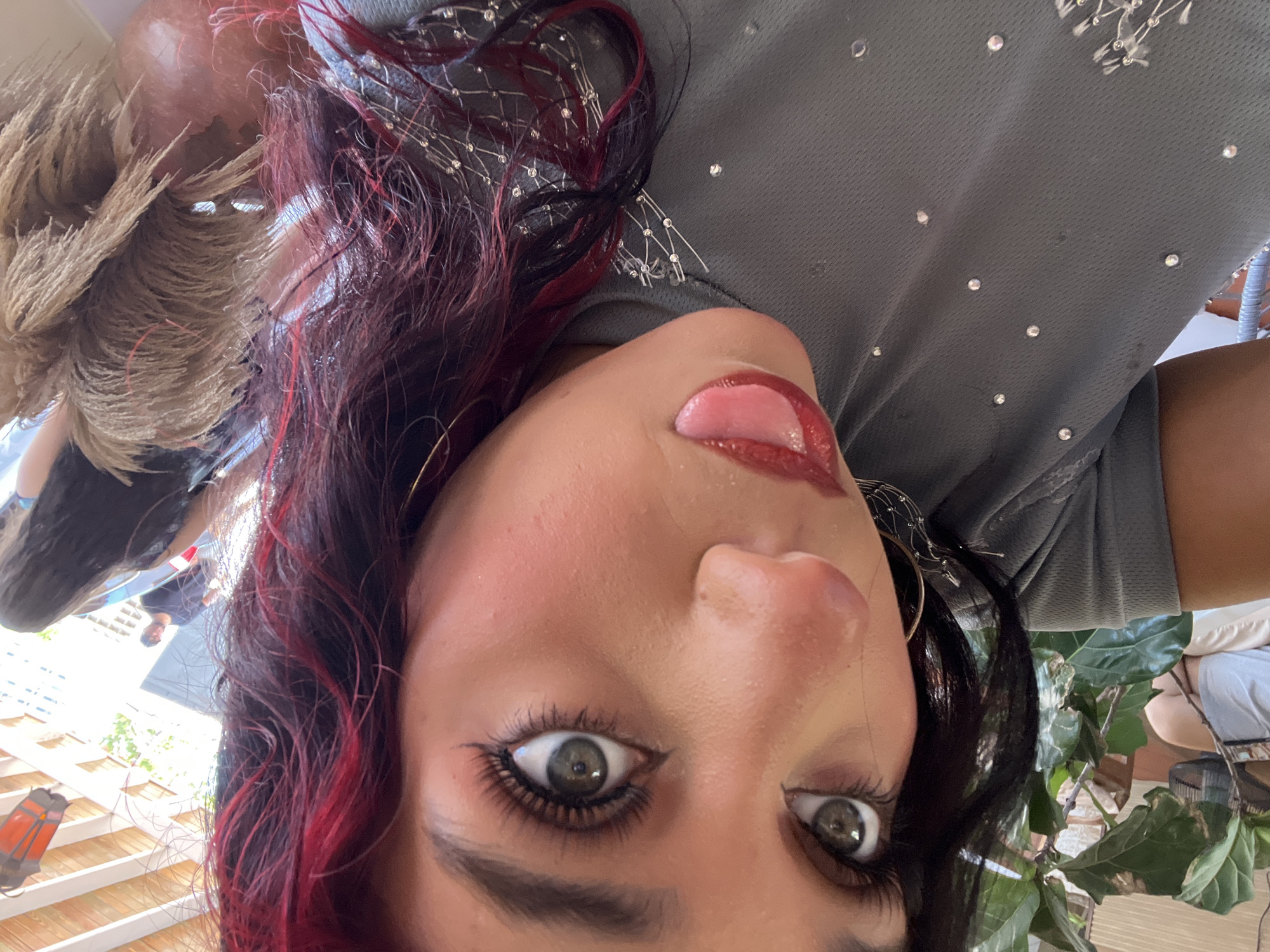 A close-up selfie shows a woman with dark hair streaked with red, her head tilted and her tongue sticking out playfully. Her eyes are wide open, and she is wearing makeup, including false eyelashes and lipstick. Her gray t-shirt is adorned with small rhinestones. In the background, a large plant is visible, as well as other people and a covered outdoor area, suggesting a casual gathering or restaurant.