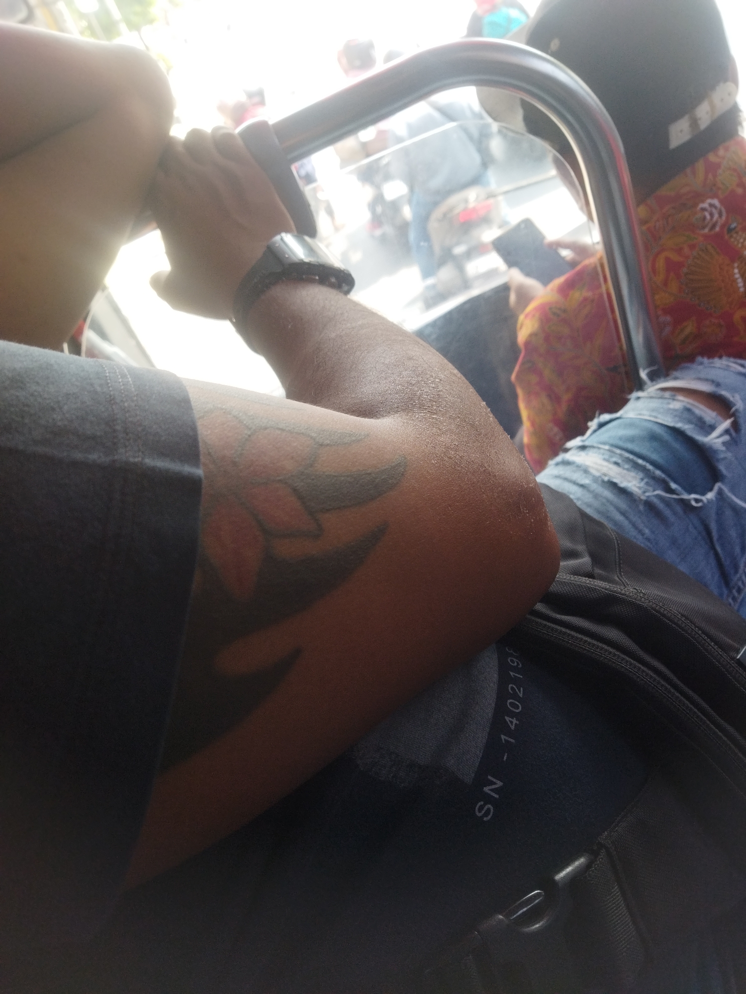 A person's arm is visible in the foreground, resting on a black backpack. The person has a dark tattoo on their arm. They are wearing a dark t-shirt. In the background, there is a curved metal bar, and beyond that, a blurred scene of a city street with people and motorcycles, suggesting the image was taken in Jombor, Indonesia. Another person in the background is wearing a red and yellow patterned shirt and ripped blue jeans, and is looking at a mobile phone. The overall lighting suggests it is daytime.