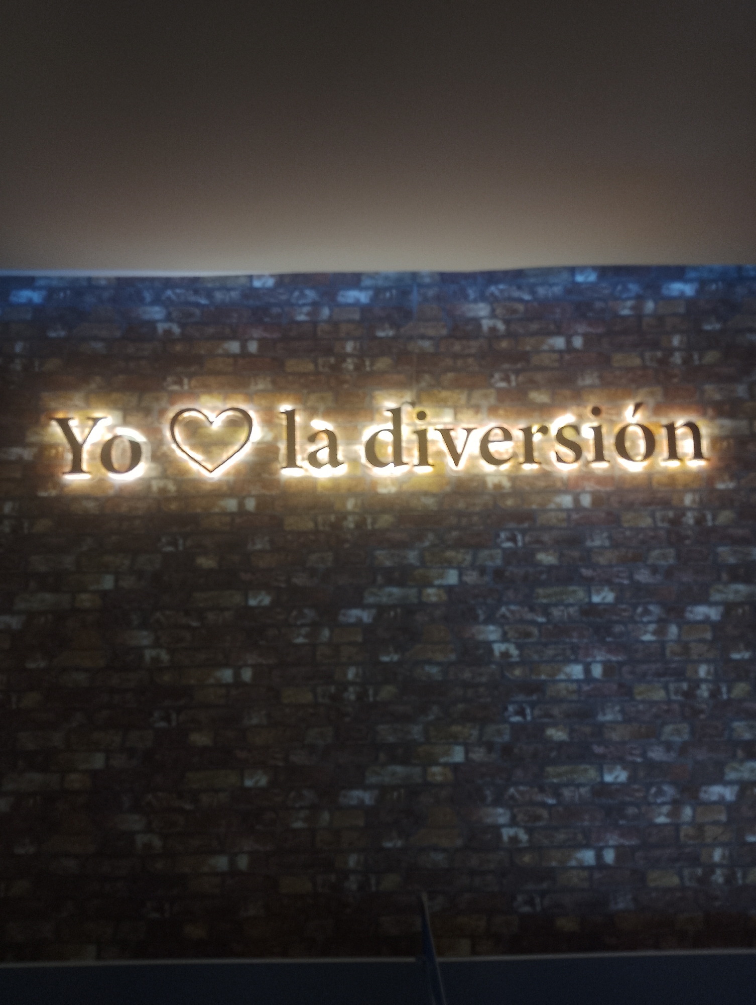 The image shows a brick wall with a sign that reads "Yo ❤️ la diversión." The sign is illuminated. The location is Porlamar, Venezuela.