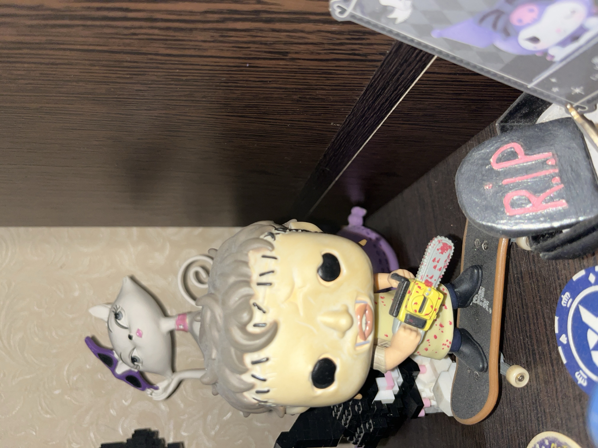 A close-up shot captures a collection of decorative figures on a shelf. In the foreground, a Funko Pop figure resembling a character from "The Texas Chain Saw Massacre" is positioned on a skateboard, holding a yellow chainsaw with a blood-splattered blade. The figure has a pale, stitched face and wears a light-colored smock. To the left of the chainsaw figure, a white cat-like figurine with large eyes and purple sunglasses sits atop a white, textured base. Behind the main figures, a dark wood-paneled shelf forms the background. In the lower right corner, a tombstone-shaped decoration with "R.I.P." in pink letters is visible. Partially obscuring the right edge of the image is a clear plastic sleeve containing a purple character with large ears, resembling a cartoon character. The lighting is dim, creating shadows that enhance the eerie aesthetic of the figures. The location is not specified in the provided information.