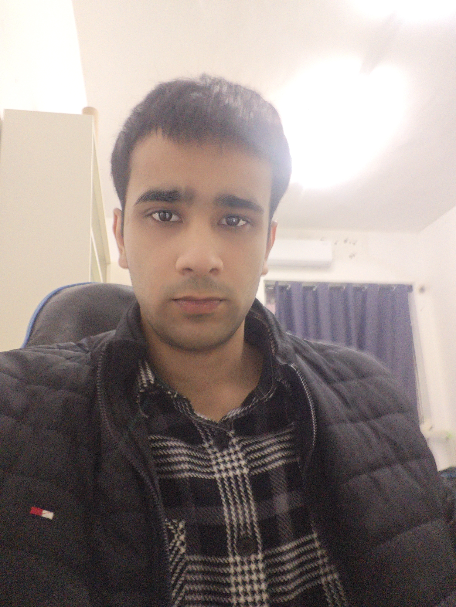 The image is a medium-shot selfie of a young man indoors. He is wearing a black quilted jacket over a black and white checkered shirt. A small rectangular patch with red and white coloring can be seen on the jacket's left side. The man has dark hair and appears to be seated in a black chair. Behind him is a wall with light-colored curtains and an air conditioning unit mounted near the ceiling. A partial view of a light-colored cabinet is visible in the upper left of the frame.
There is no information within the image to indicate the location is Chongqing High-tech Zone, China.